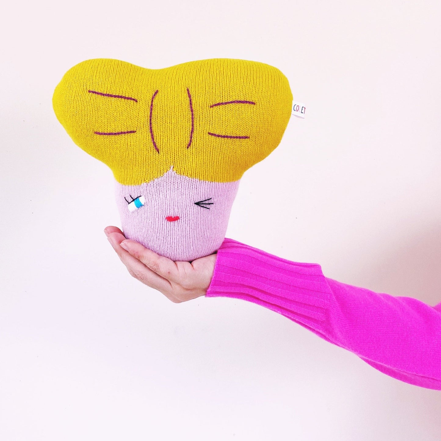 Hand holding a pink and yellow plush toy with a face on a white background