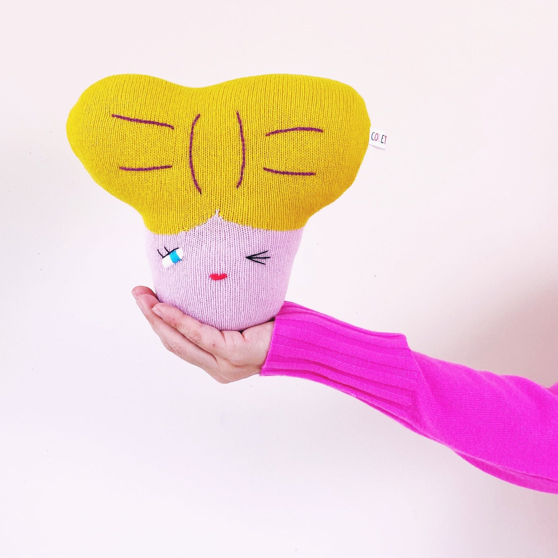 Hand holding a pink and yellow plush toy with a face on a white background