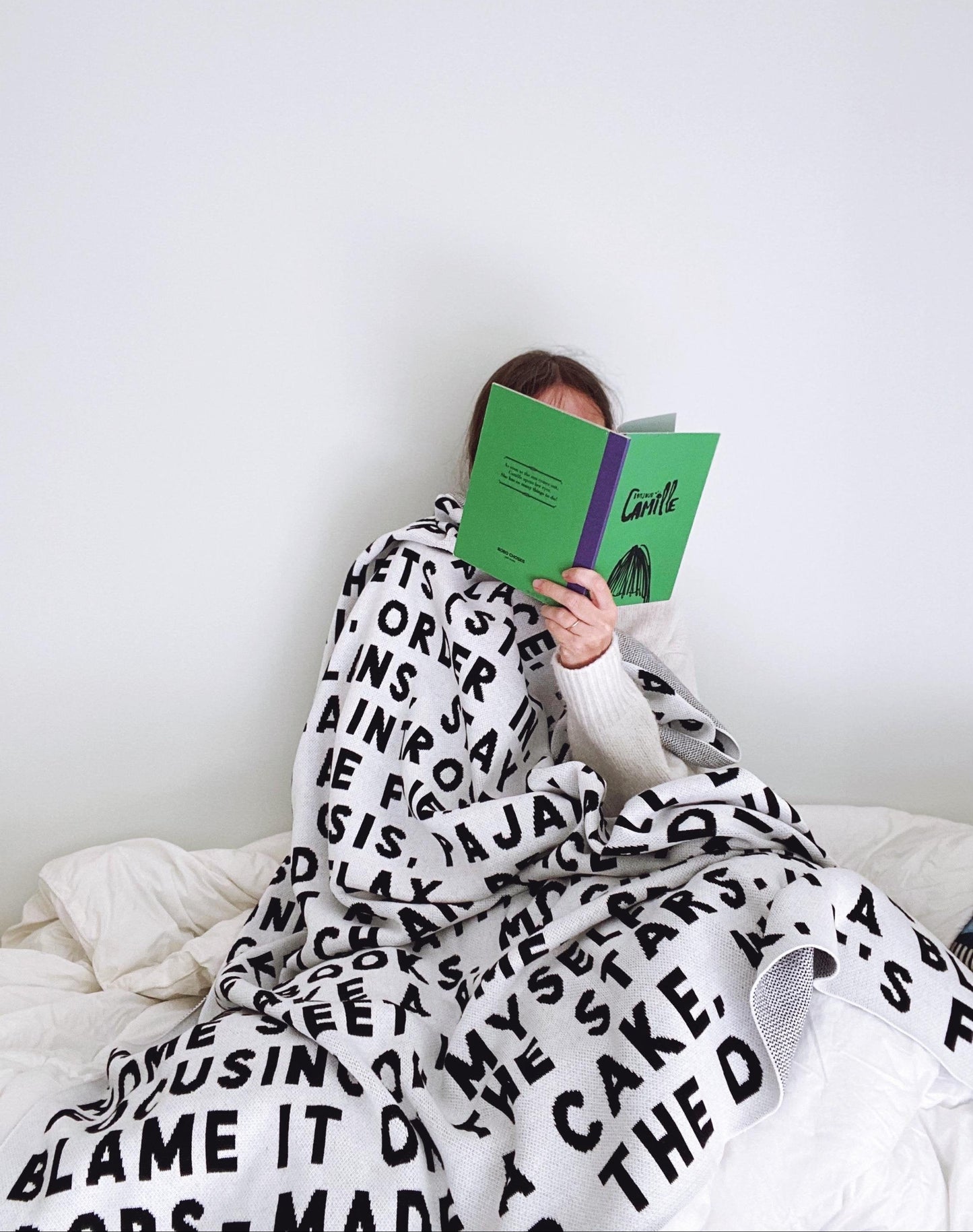 Person reading a book under a blanket with text, sitting on a bed.