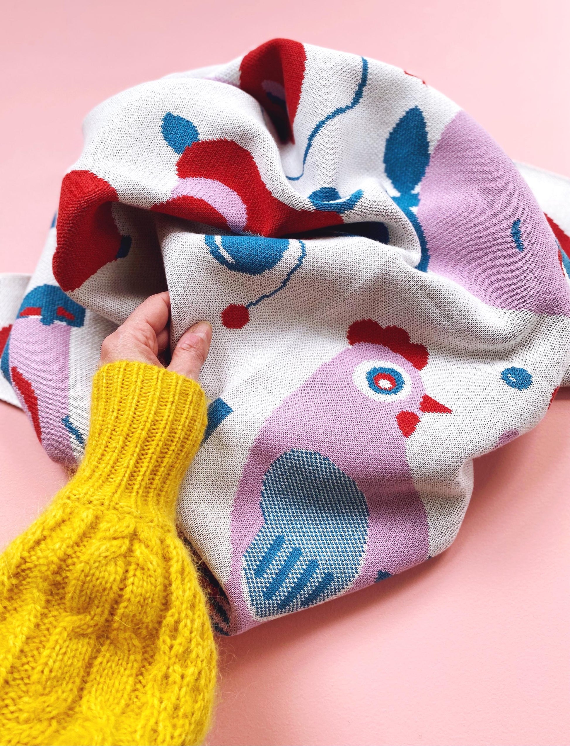Colorful knitted blanket with chicken pattern held by a hand in yellow sleeve against a pink background