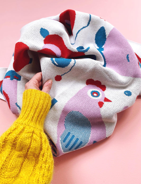 Colorful knitted blanket with chicken pattern held by a hand in yellow sleeve against a pink background