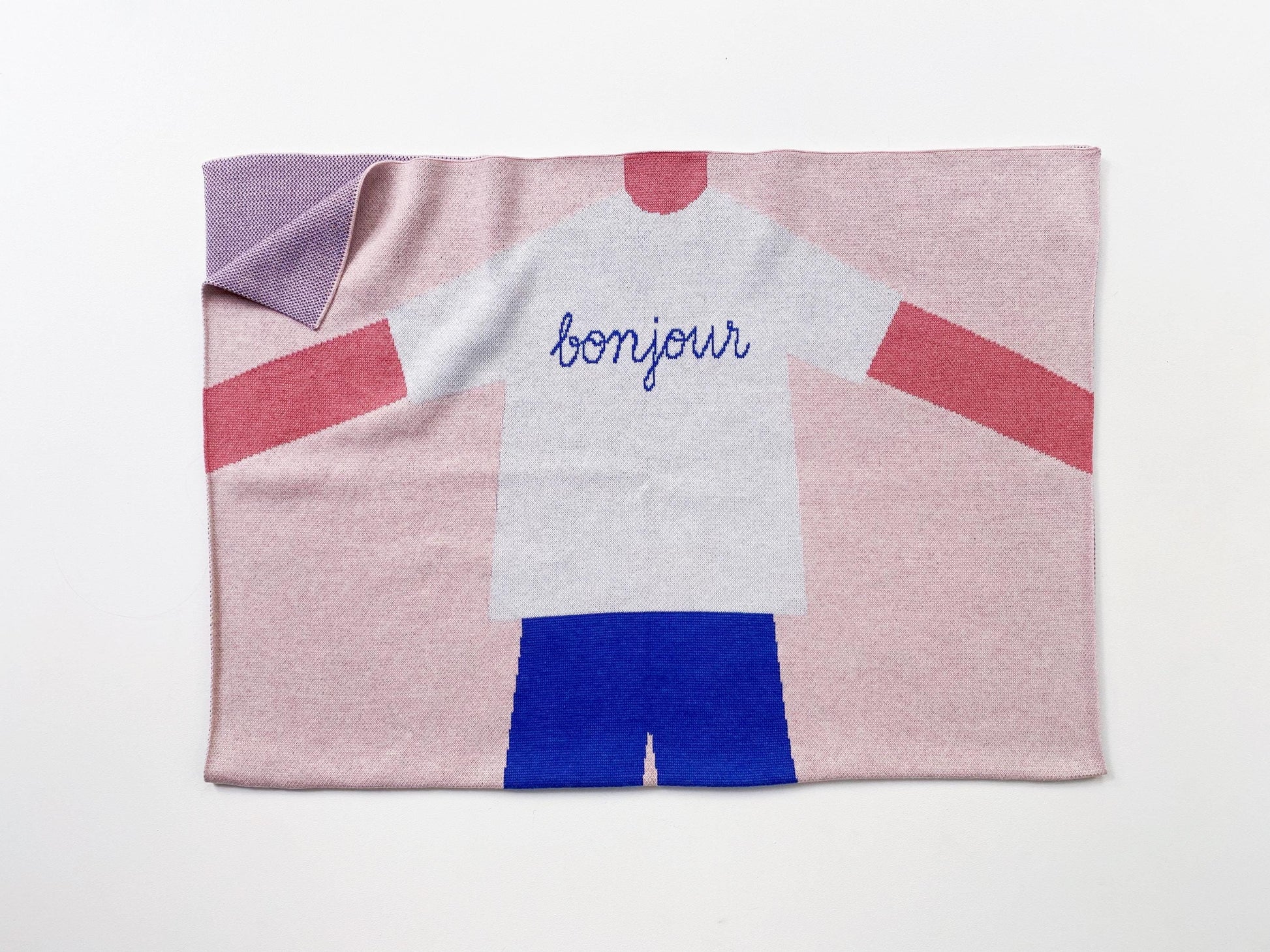 Knit baby blanket with a design of a person wearing a shirt and shorts, with 'bonjour' text on a pink background.