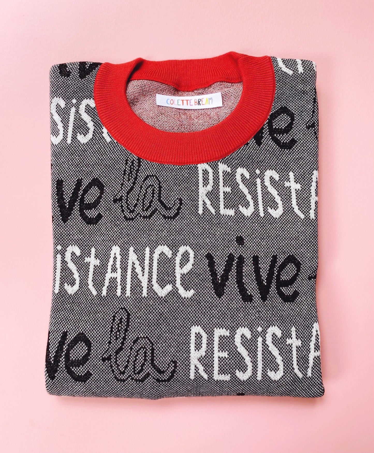 Folded black and white sweater with red neckline and 'Vive la Resistance' text on a pink background