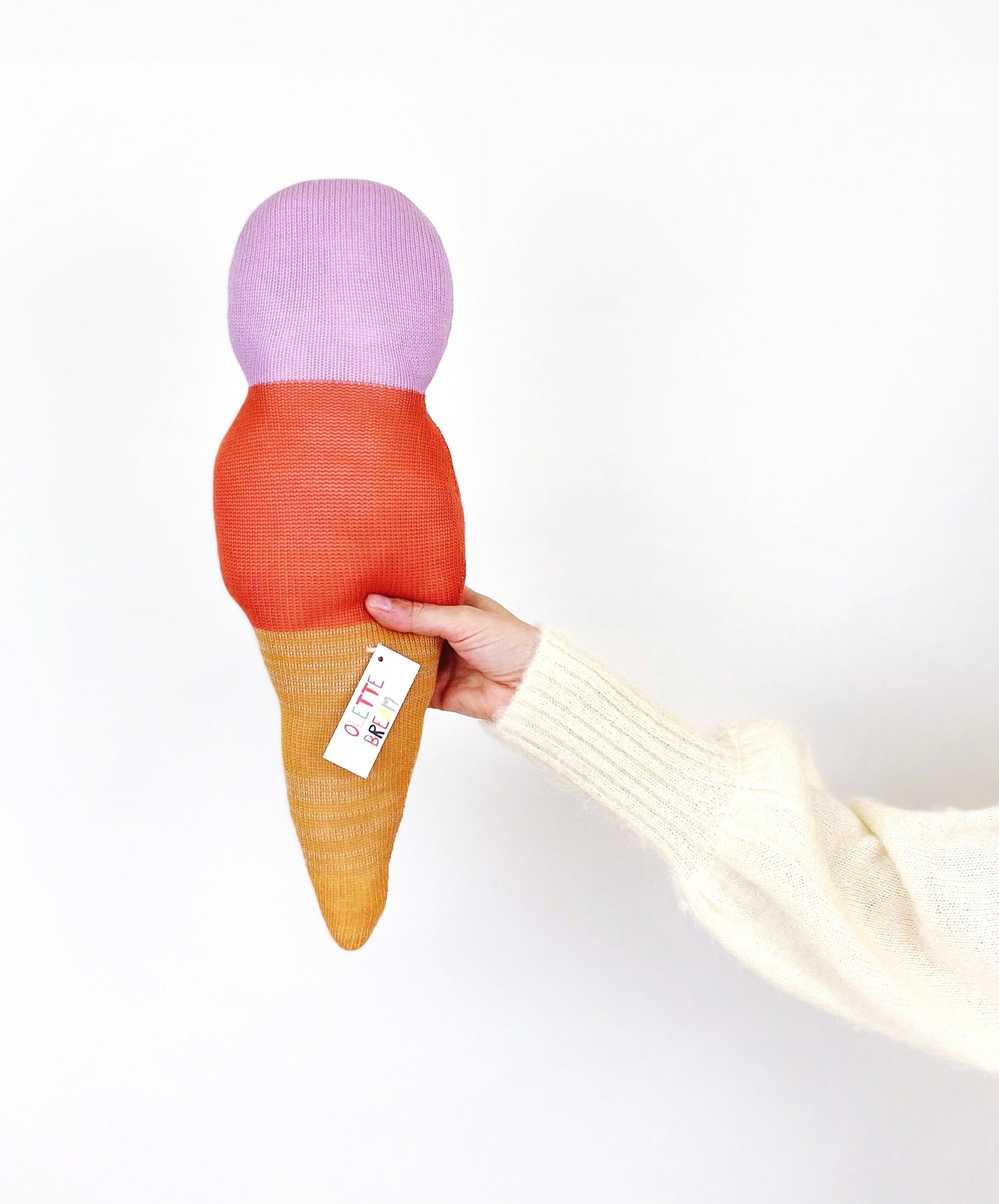 Hand holding a plush ice cream cone toy with a purple top, red middle, and yellow bottom on a white background.