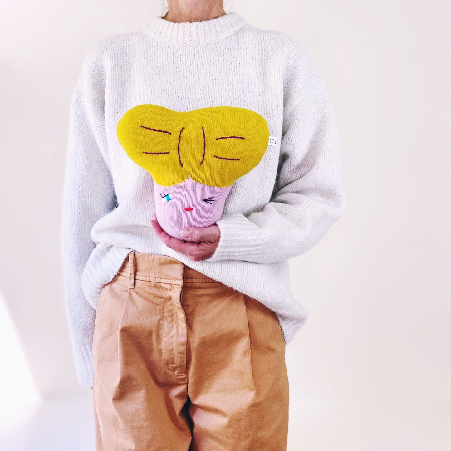 Person wearing a white sweater holding a colorful plush toy character design, holding a pink plush toy.