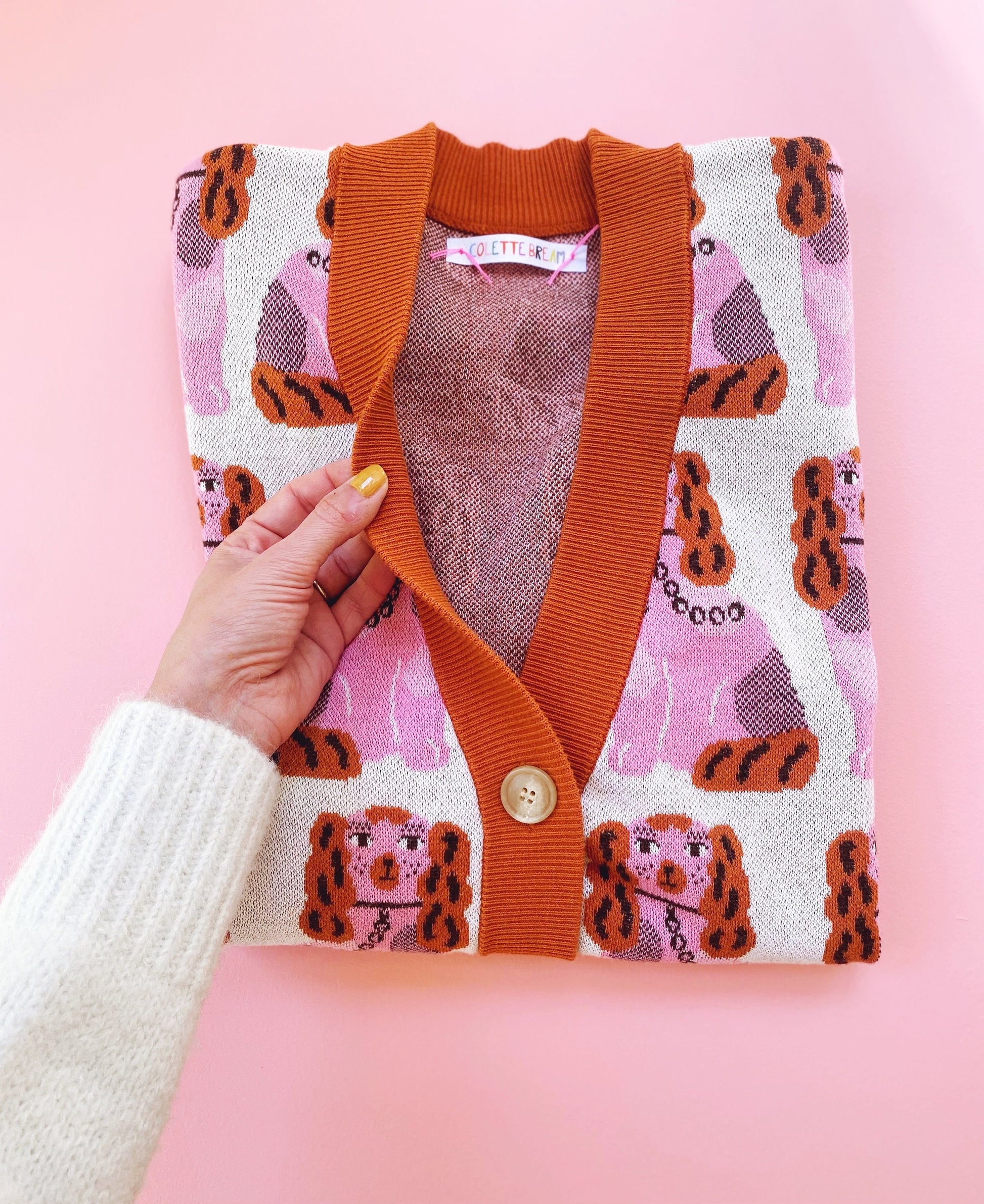 Hand holding a patterned cardigan with orange trim against a pink background