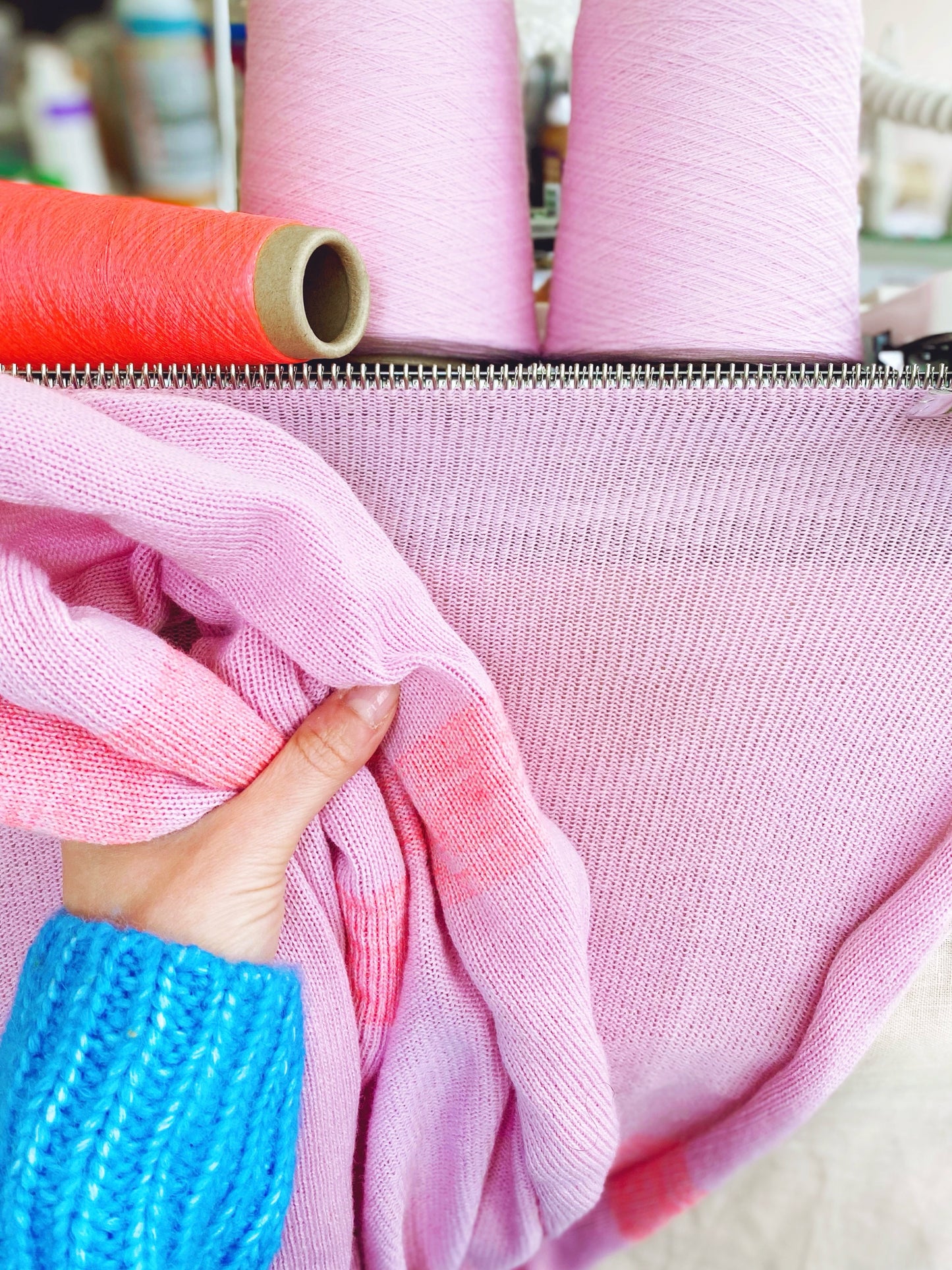 Close-up of pink fabric with a hand holding it, with spools of thread in the background.