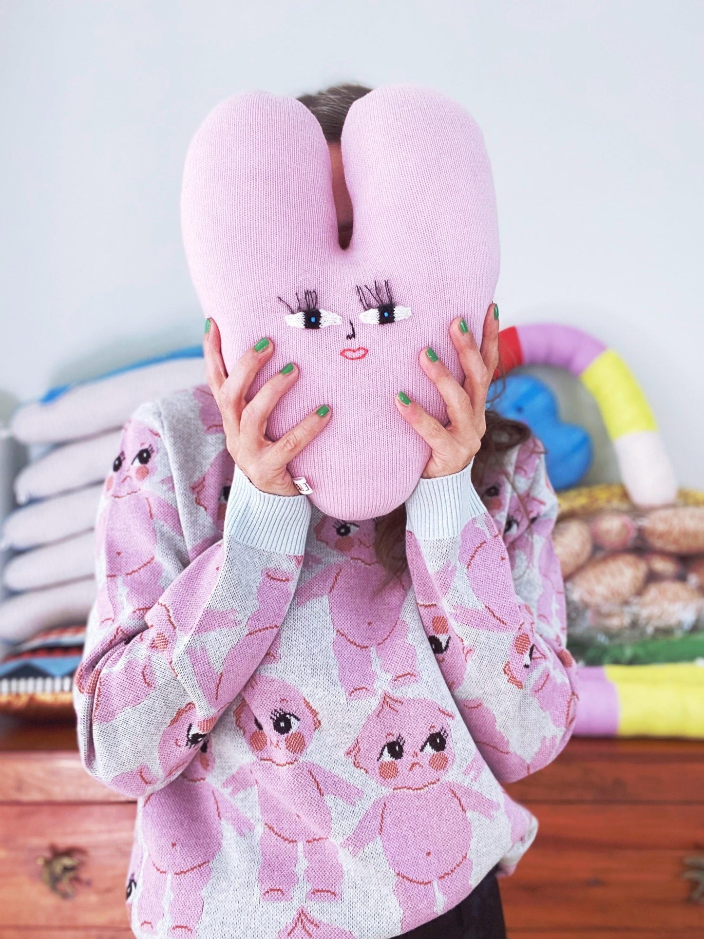 Person wearing a pink sweater with cartoon character design, holding a pink heart shaped plush toy with face design