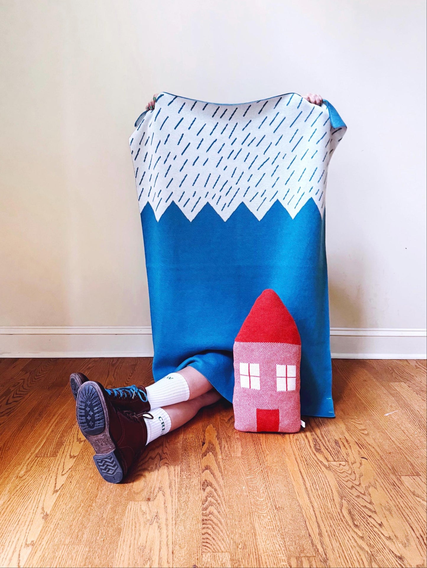 Person sitting on a wooden floor with a blue and white mountain patterned blanket and a red house-shaped pillow.