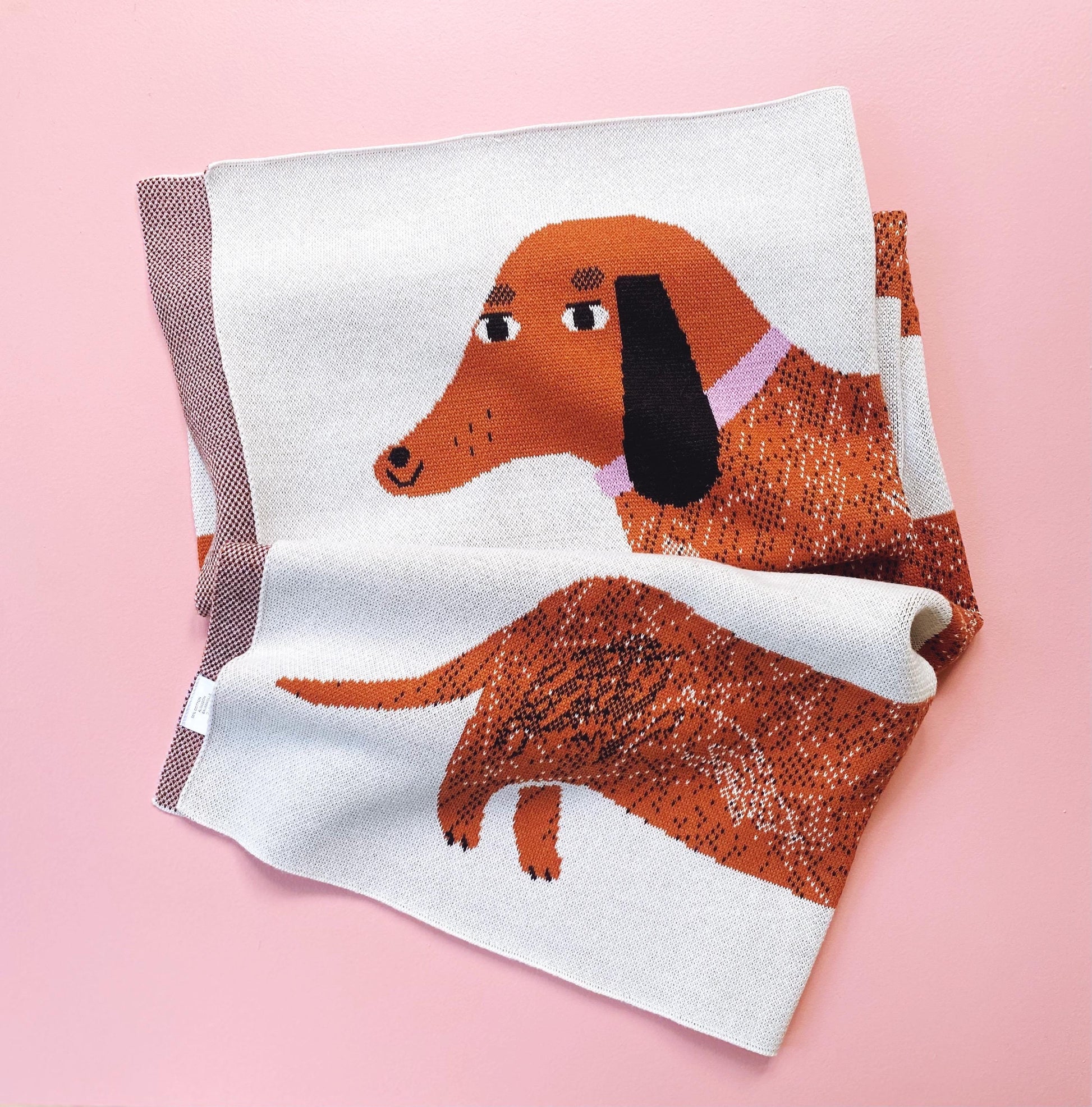 Folded knit blanket with a brown dachshund design on a pink background
