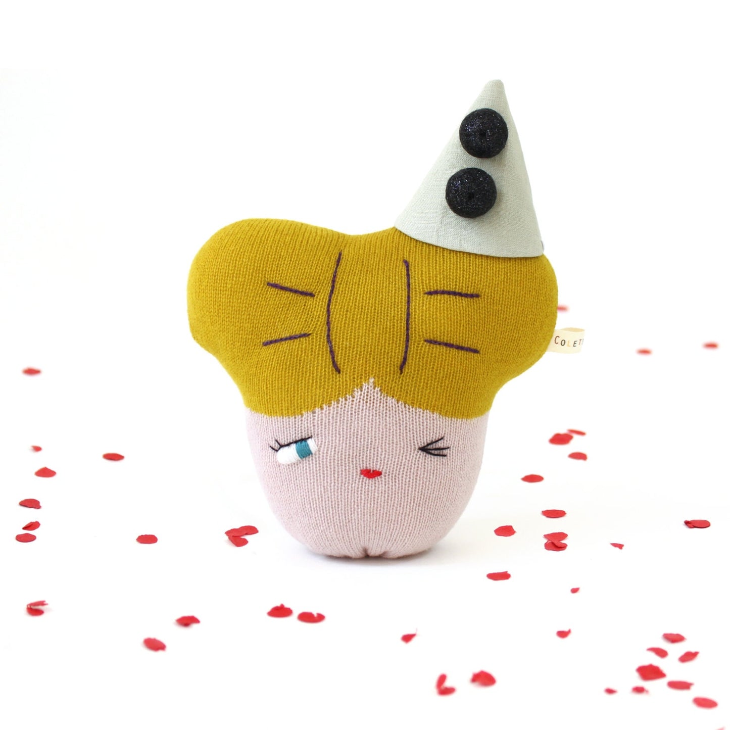 Toy resembling a face with a yellow hair and pink face, surrounded by red heart shaped confetti on a white background