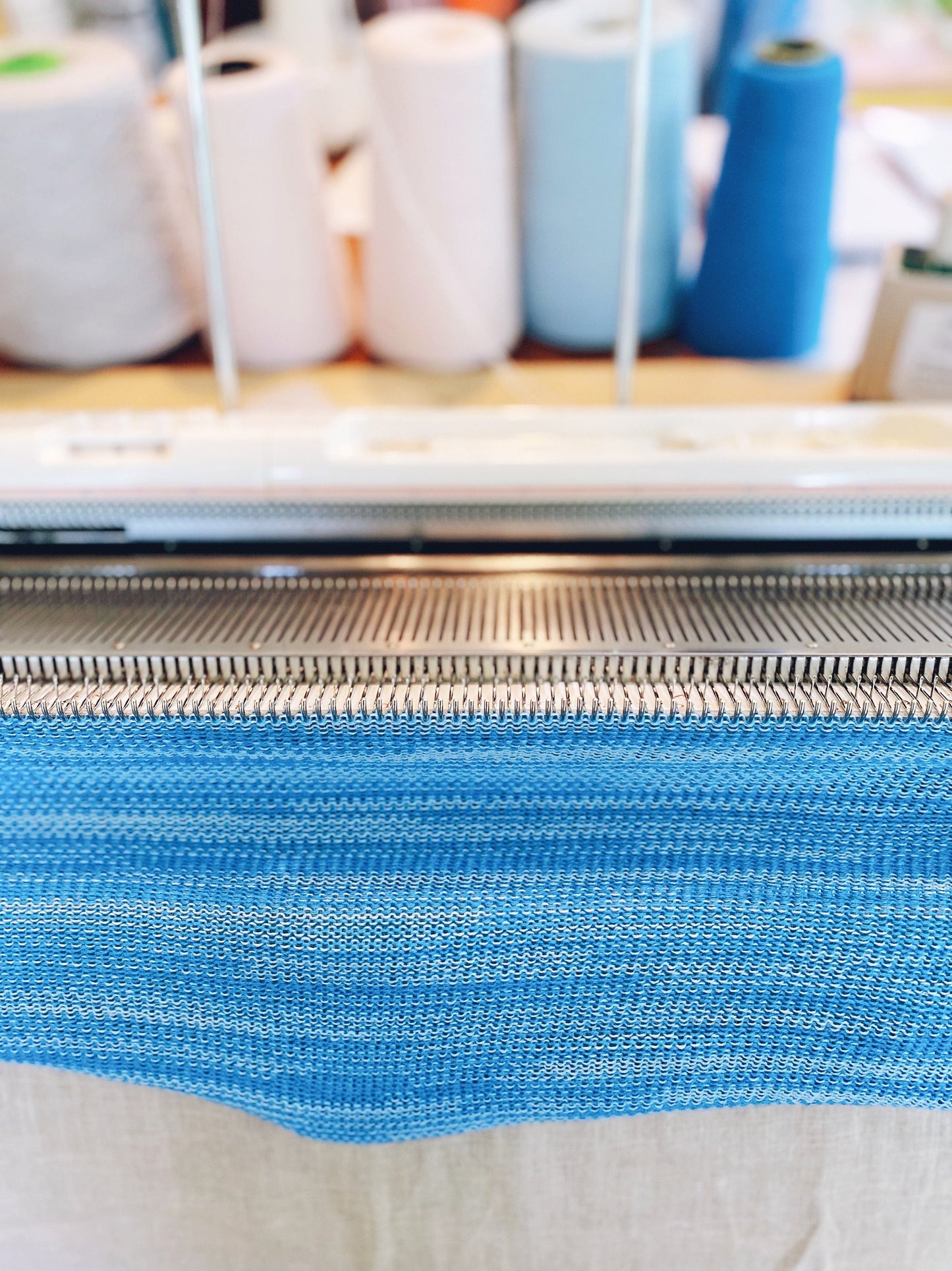 Close-up of blue fabric being knitted on a machine with spools of thread in the background.