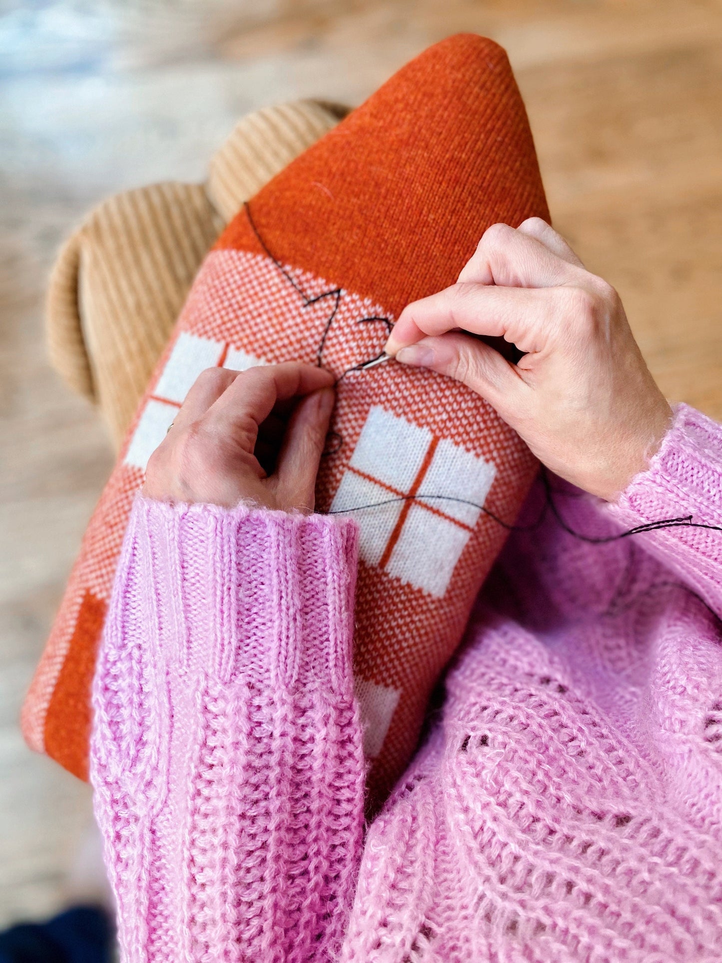 Person sewing a red fabric item with white patterns, wearing a pink sweater.