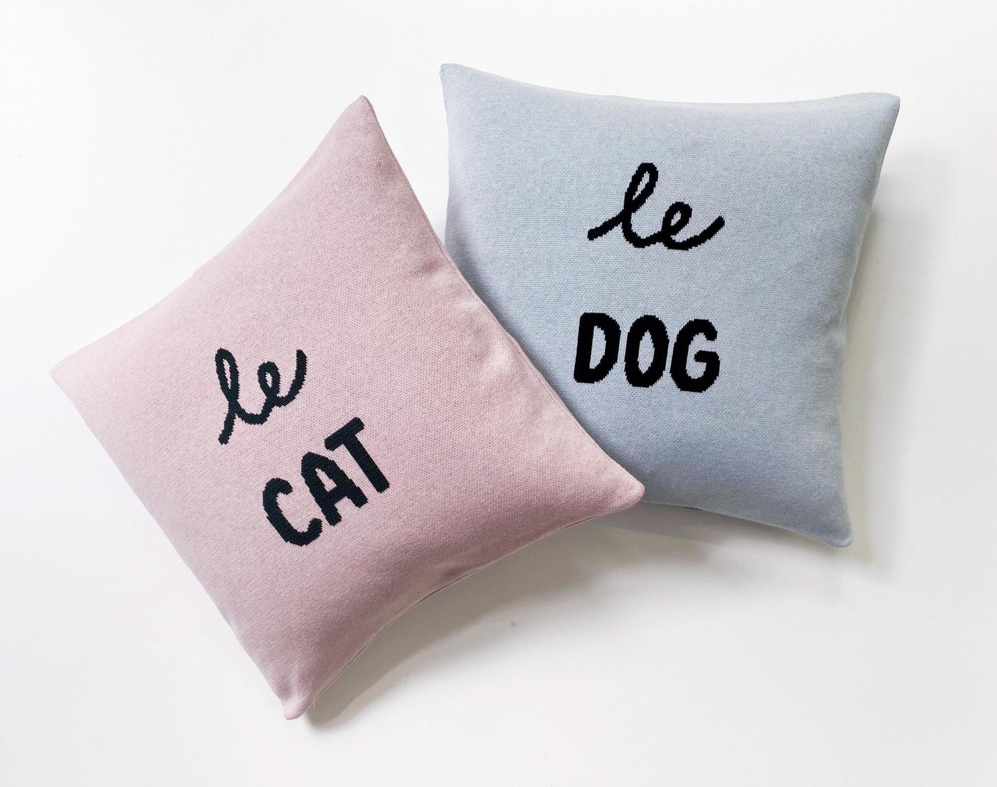 Two knitted cushions, one pink and one gray, with 'le cat' and 'le dog' text on a white background.