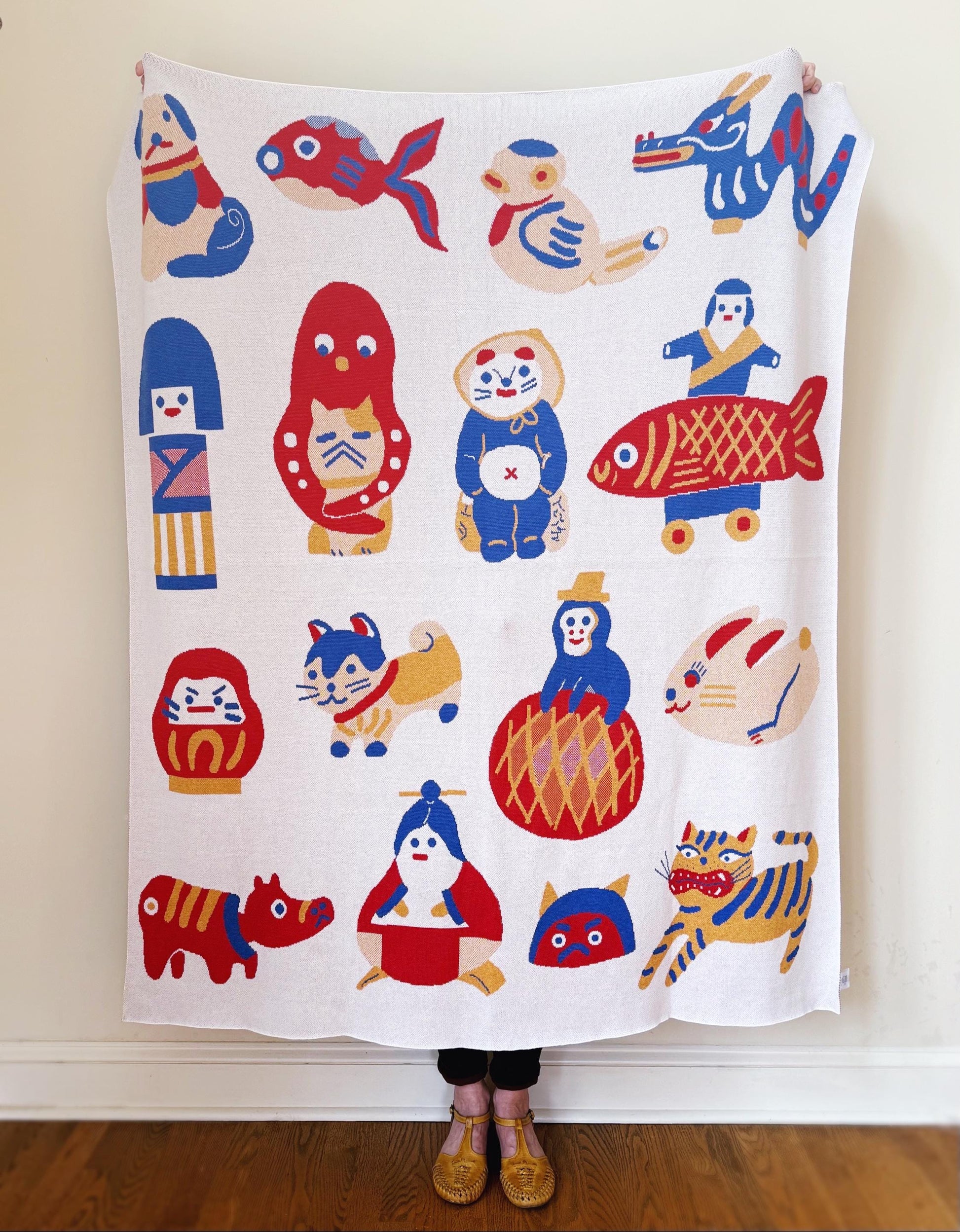Person holding a colorful knit blanket with cartoon japanese folk toys animal designs against a plain wall.