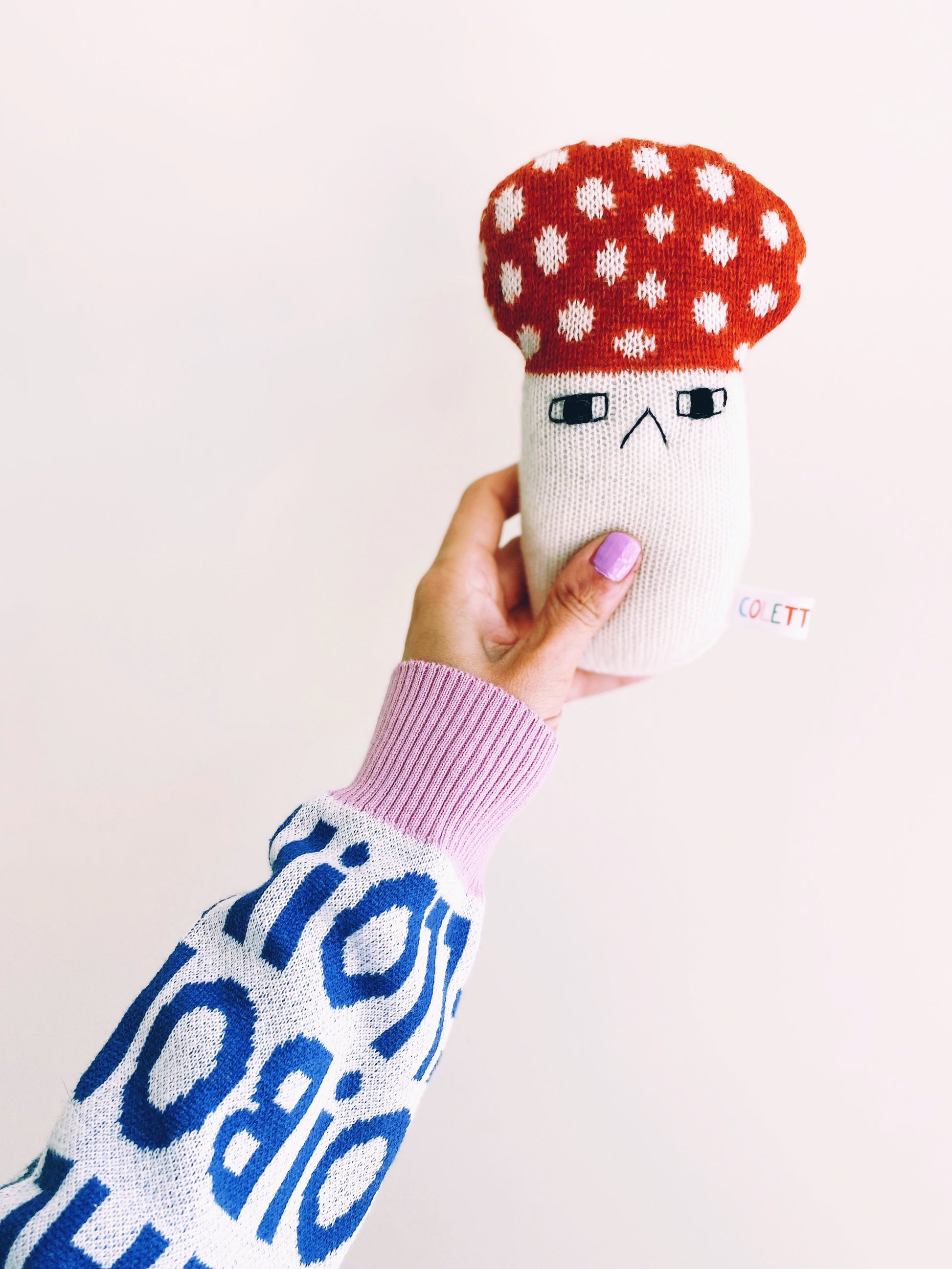 Soft knitted baby rattle toy with a mushroom design held by a person wearing a colorful sweater.