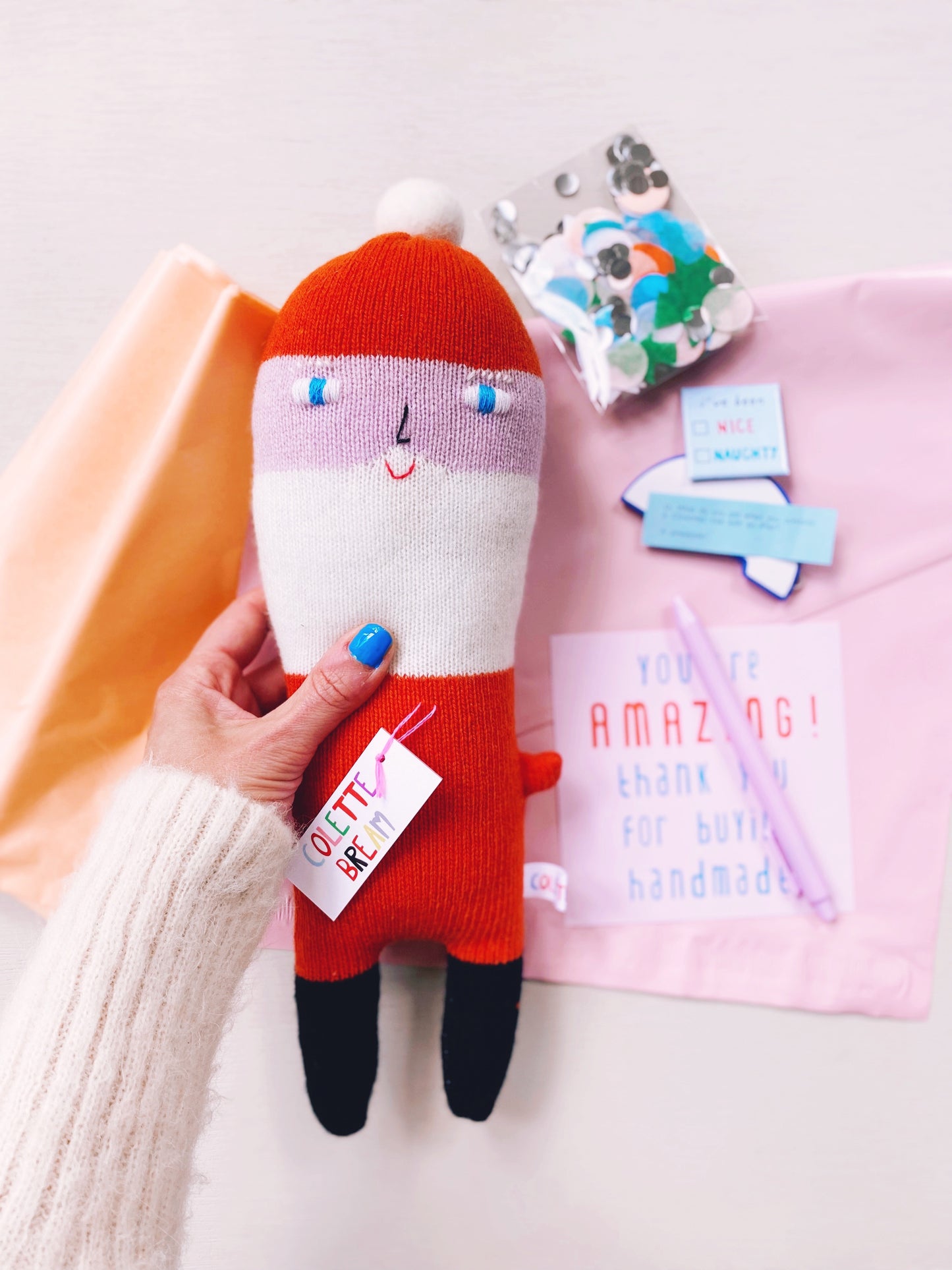 Handmade Santa plush doll held by a person with a pink background and craft materials.