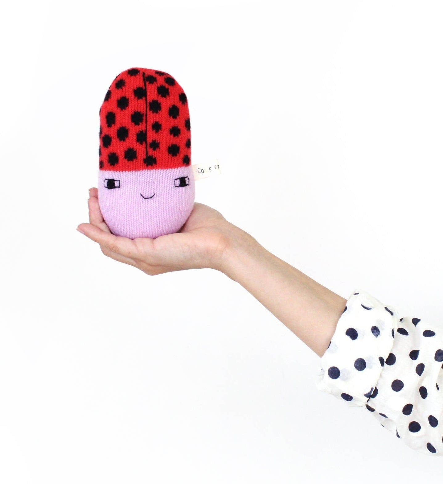 Hand holding a soft knitted baby rattle plush toy shaped like a ladybug with a red and black dots on a white background