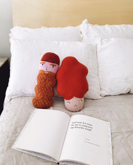 Two knitted dolls with red hair and a beard on a bed next to an open book.