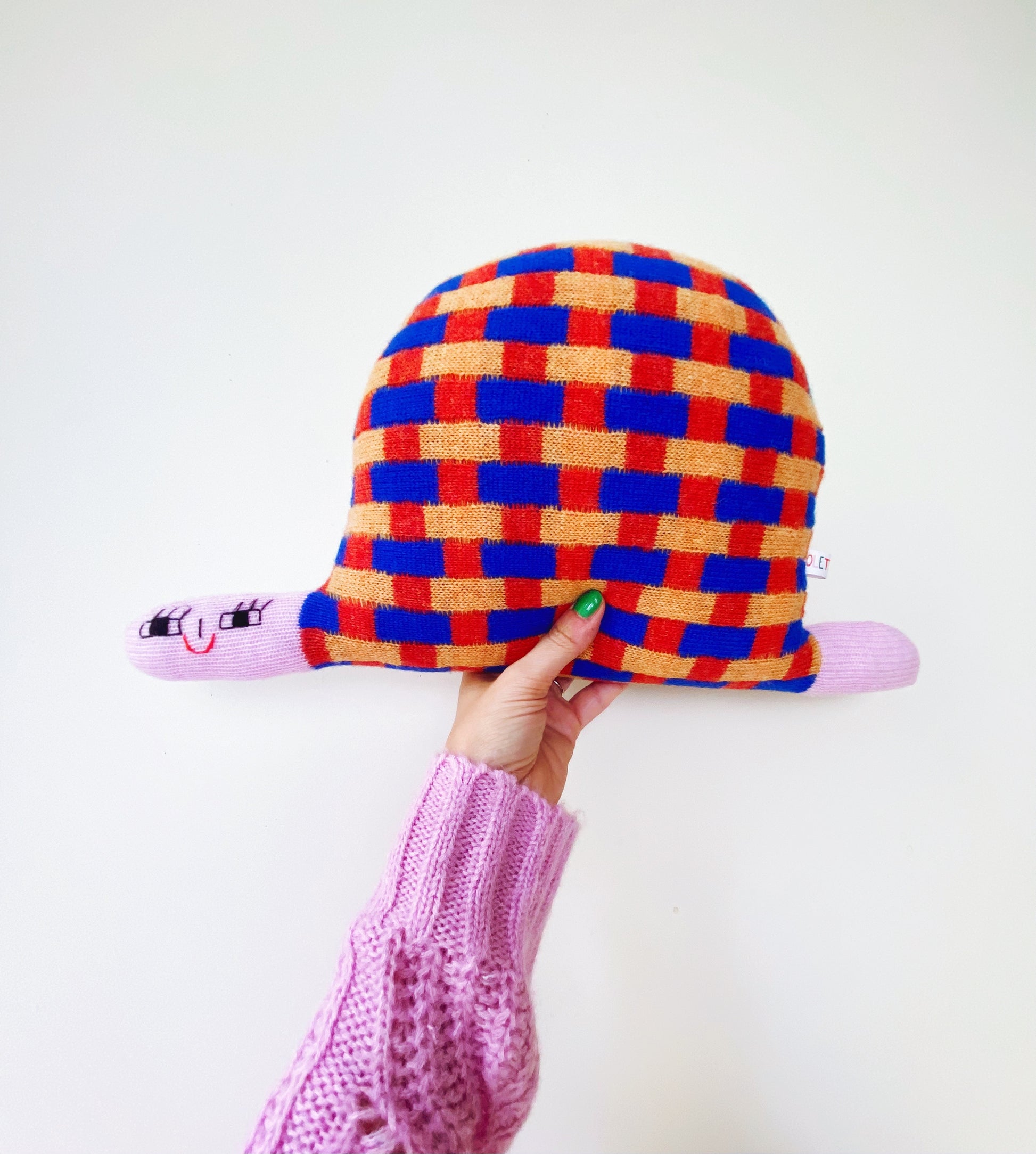 Colorful checkered soft knitted turtle plush toy held by a person wearing a pink sweater against a white background