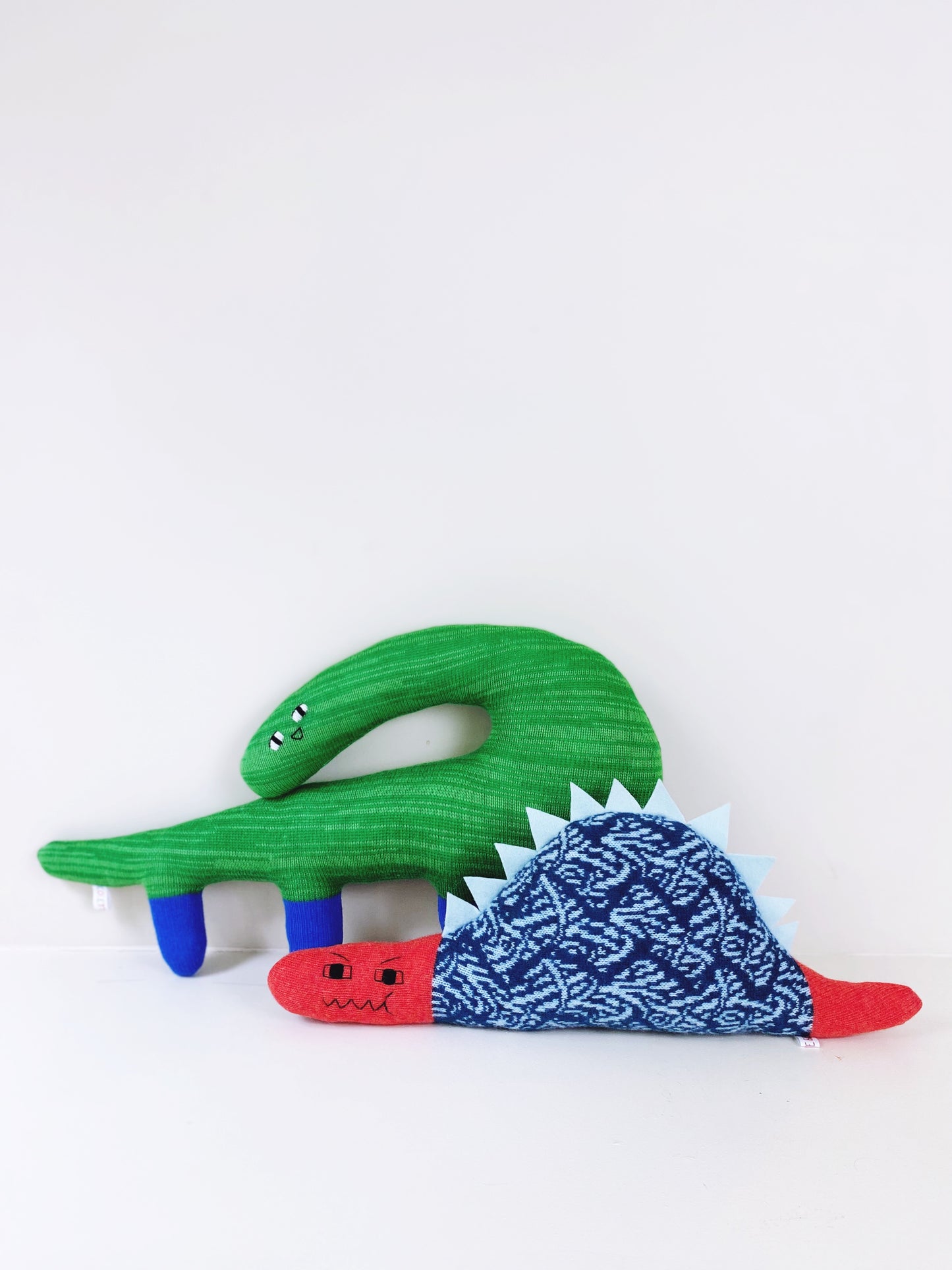 Two toy dinosaur cushions, one green and one red with a blue patterned body, on a white background.