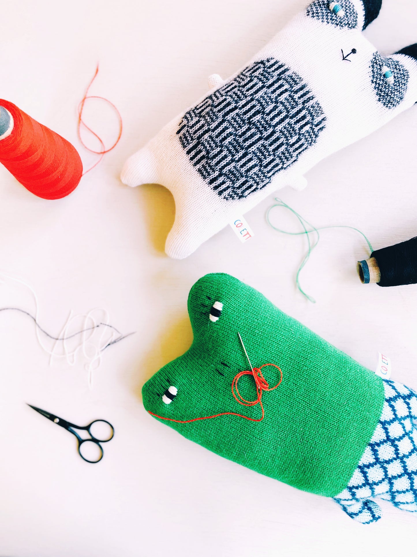Green knitted frog plush toy with red thread, scissors, and yarn on a white background