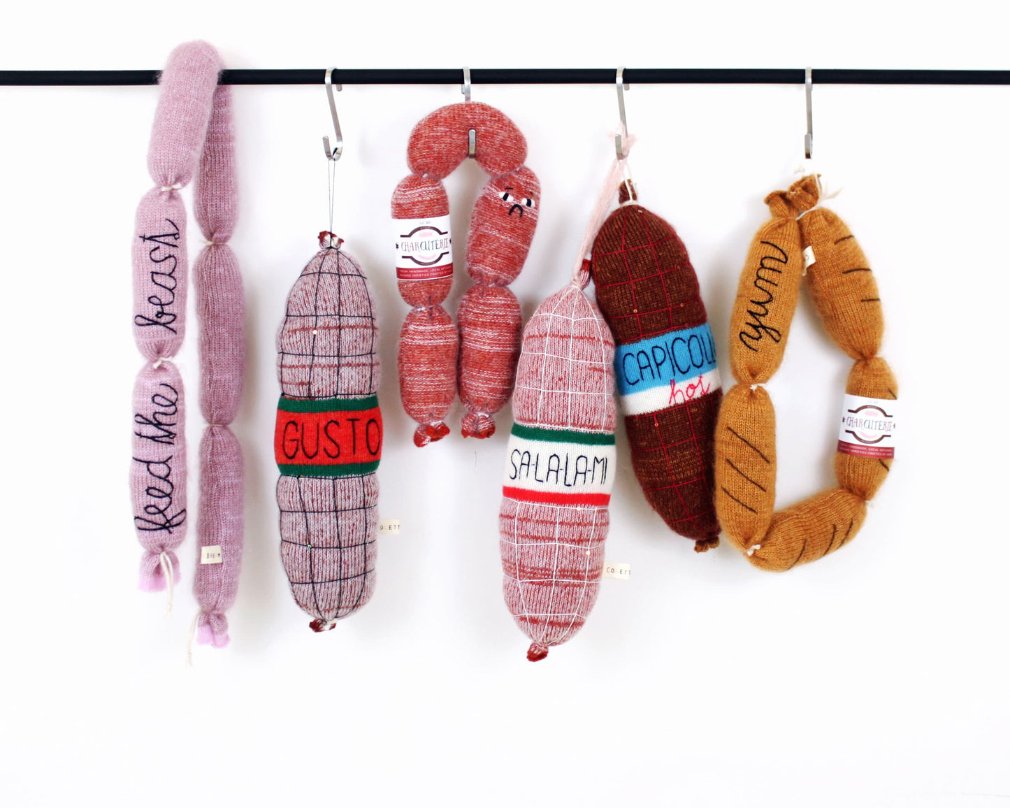 Plush toy knitted sausages and salami hanging on a rack with various labels.