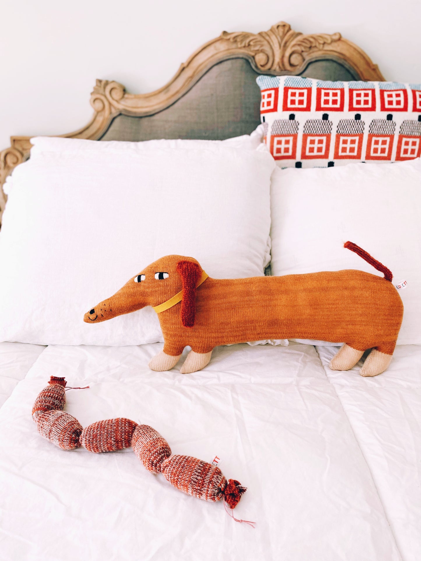 Knitted dachshund-shaped pillow plush with a knitted sausage on a bed with decorative headboard and pillowcase.