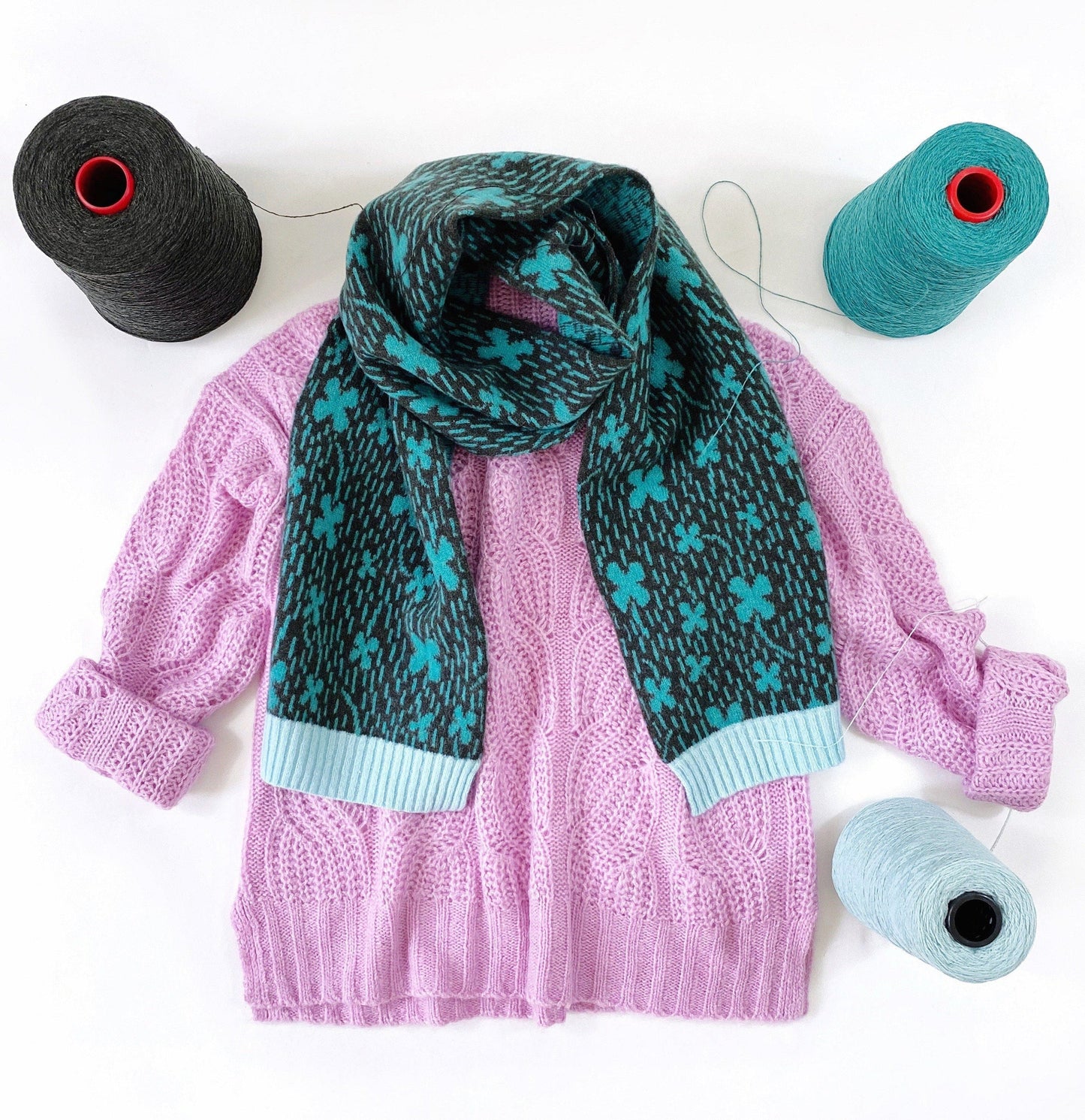Pink sweater with a green and blue patterned four-leaf clover scarf, surrounded by yarn on a white background