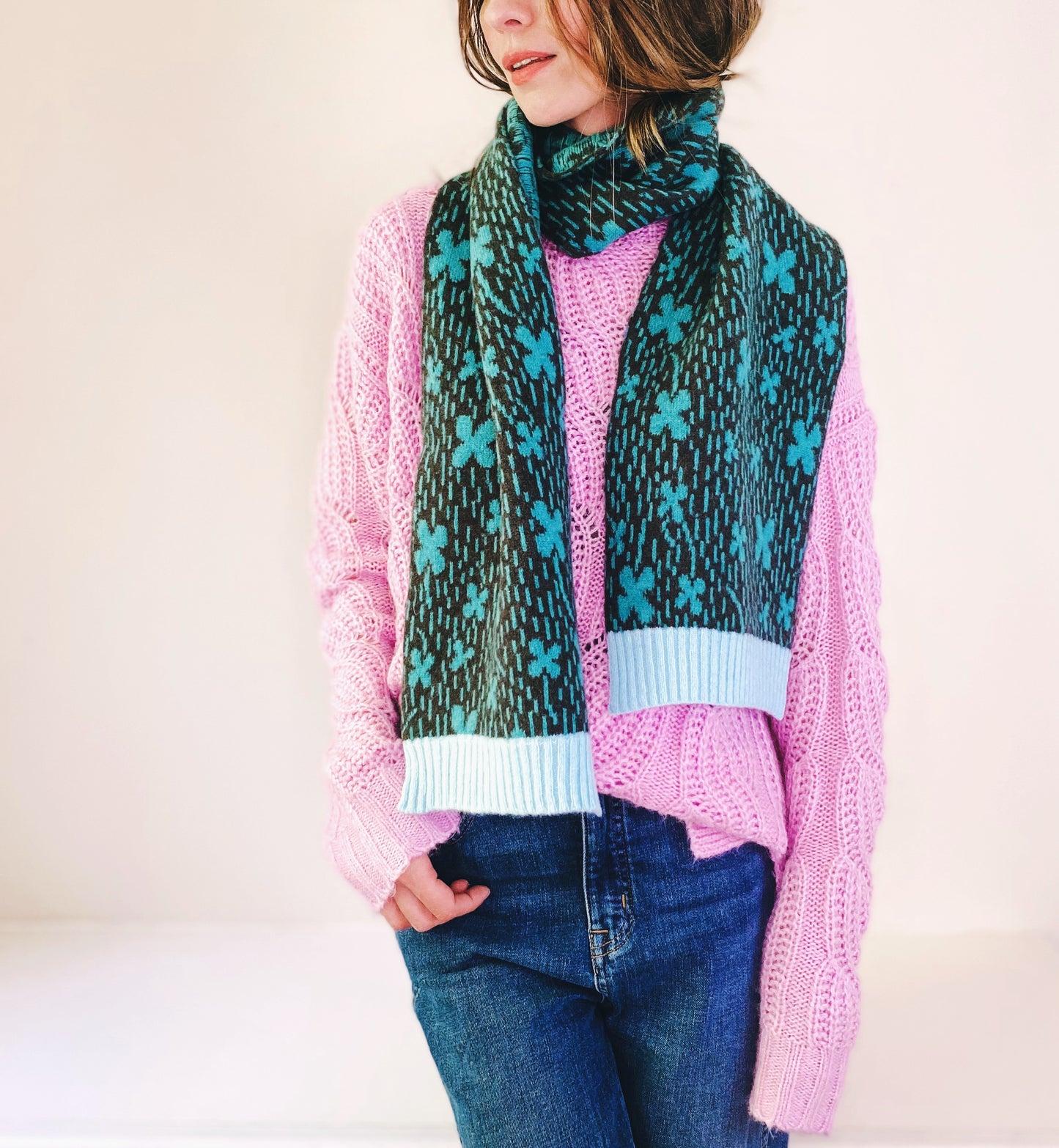 Person wearing a pink sweater, blue jeans, and a patterned knitted shamrock scarf against a white background
