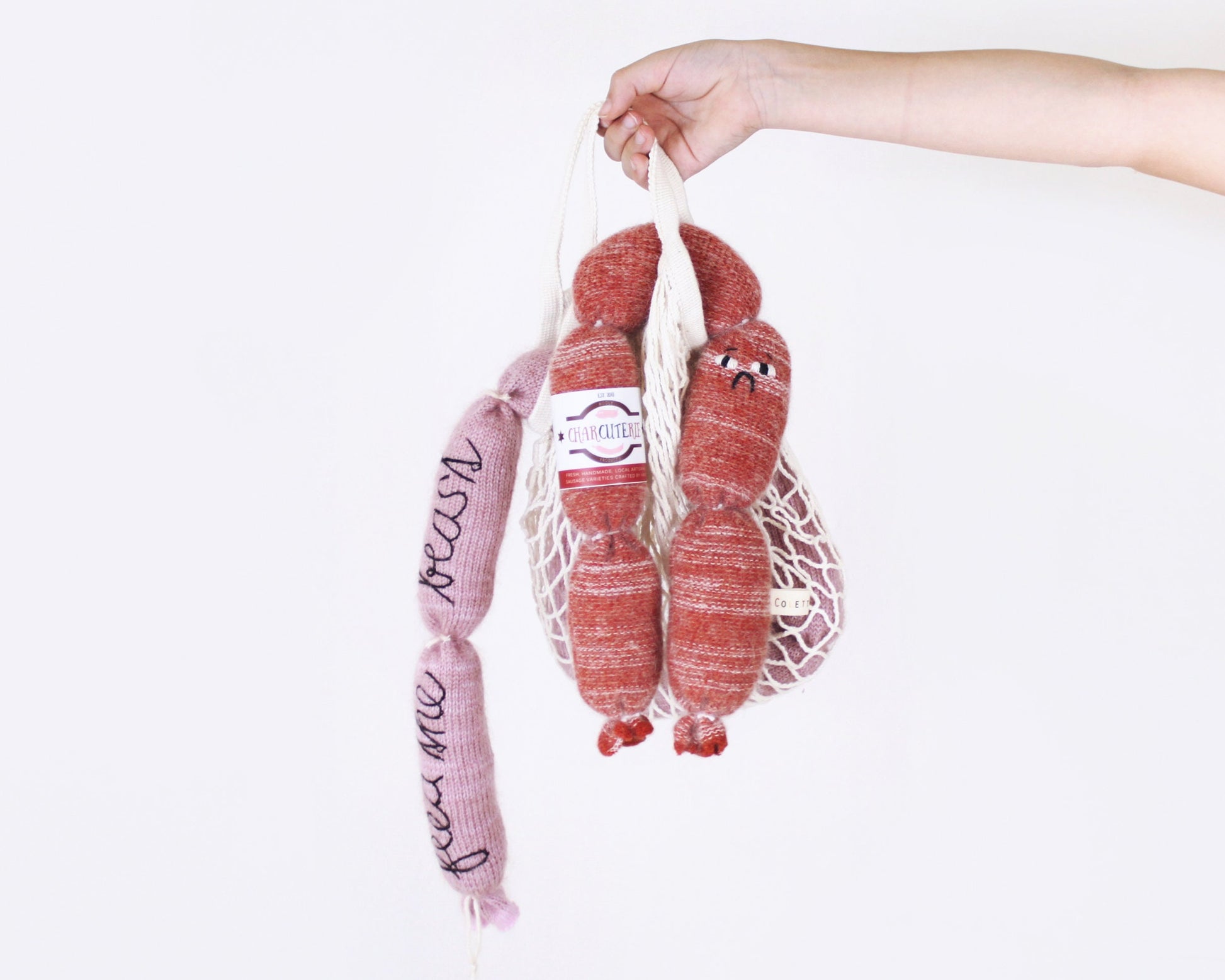 Hand holding a mesh bag with pink and red knitted plush toy sausage links against a white background