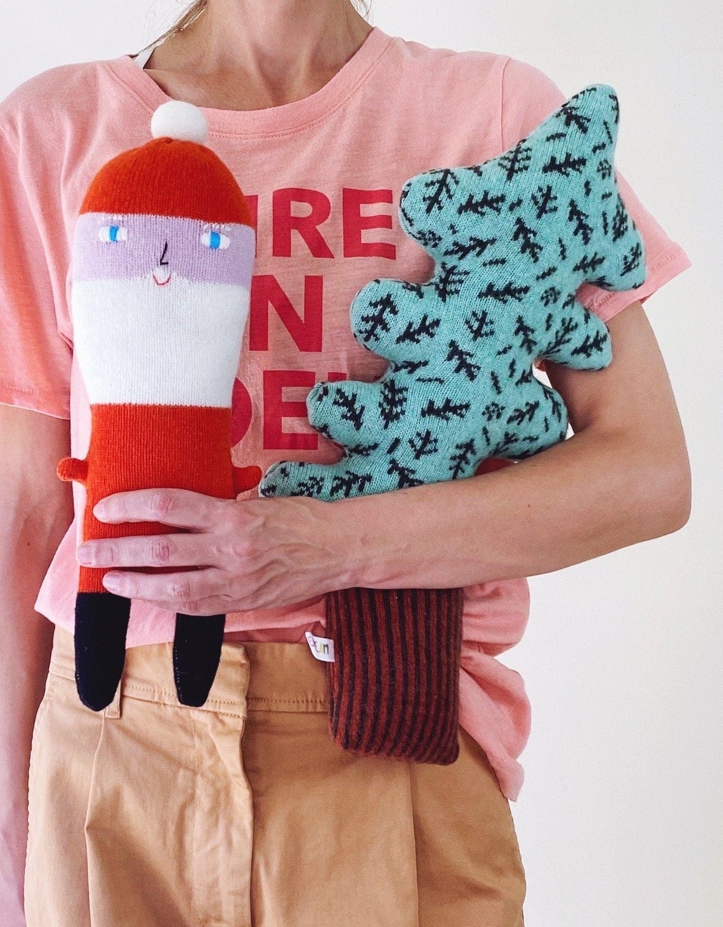 Person holding two knitted toys, one red and white santa claus, one green tree pillow, against a pink shirt background.