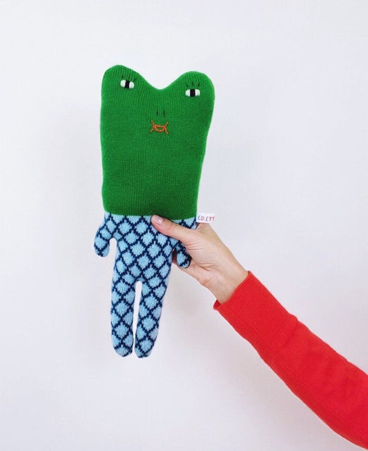 Green and blue plush toy frog with a face design held by a hand on a white background