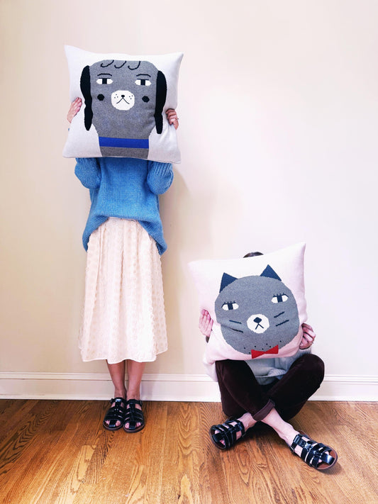 Two people holding cat and dog themed cushions against a plain wall.