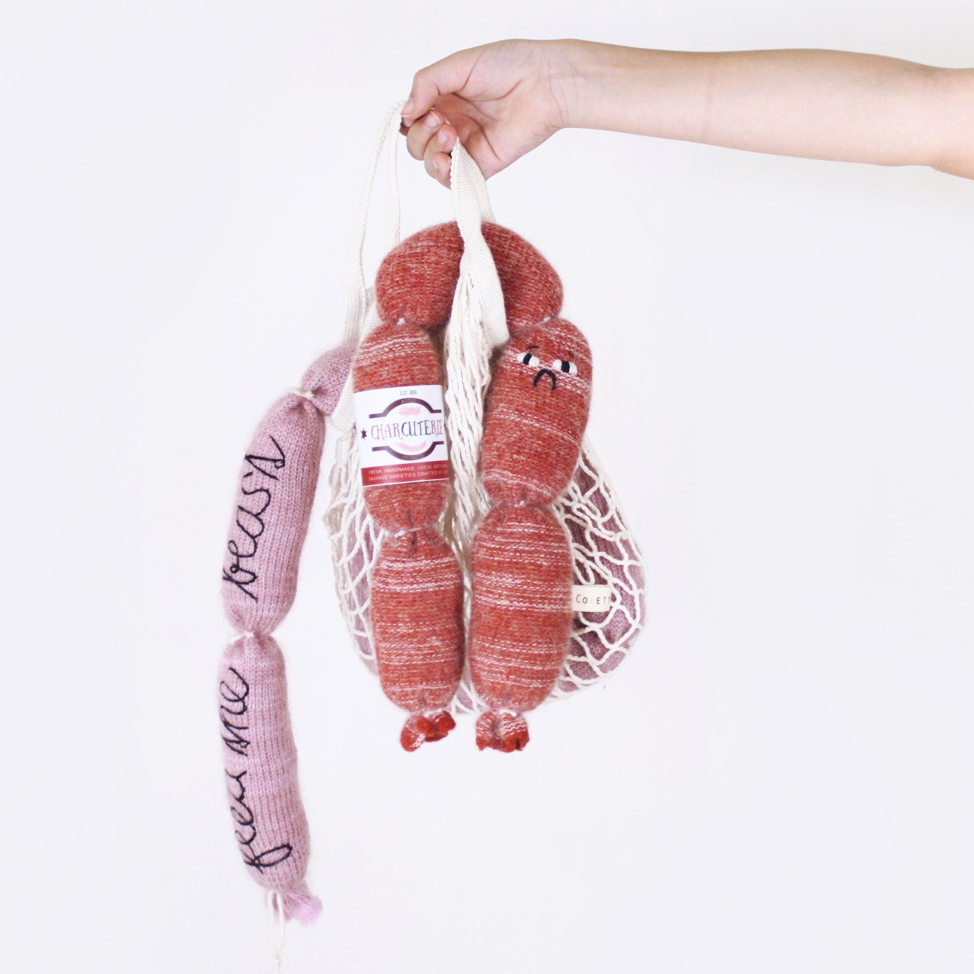 Hand holding a mesh bag with pink and red knitted sausages plush toys against a white background