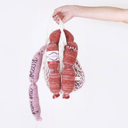 Hand holding a mesh bag with pink and red knitted sausages plush toys against a white background