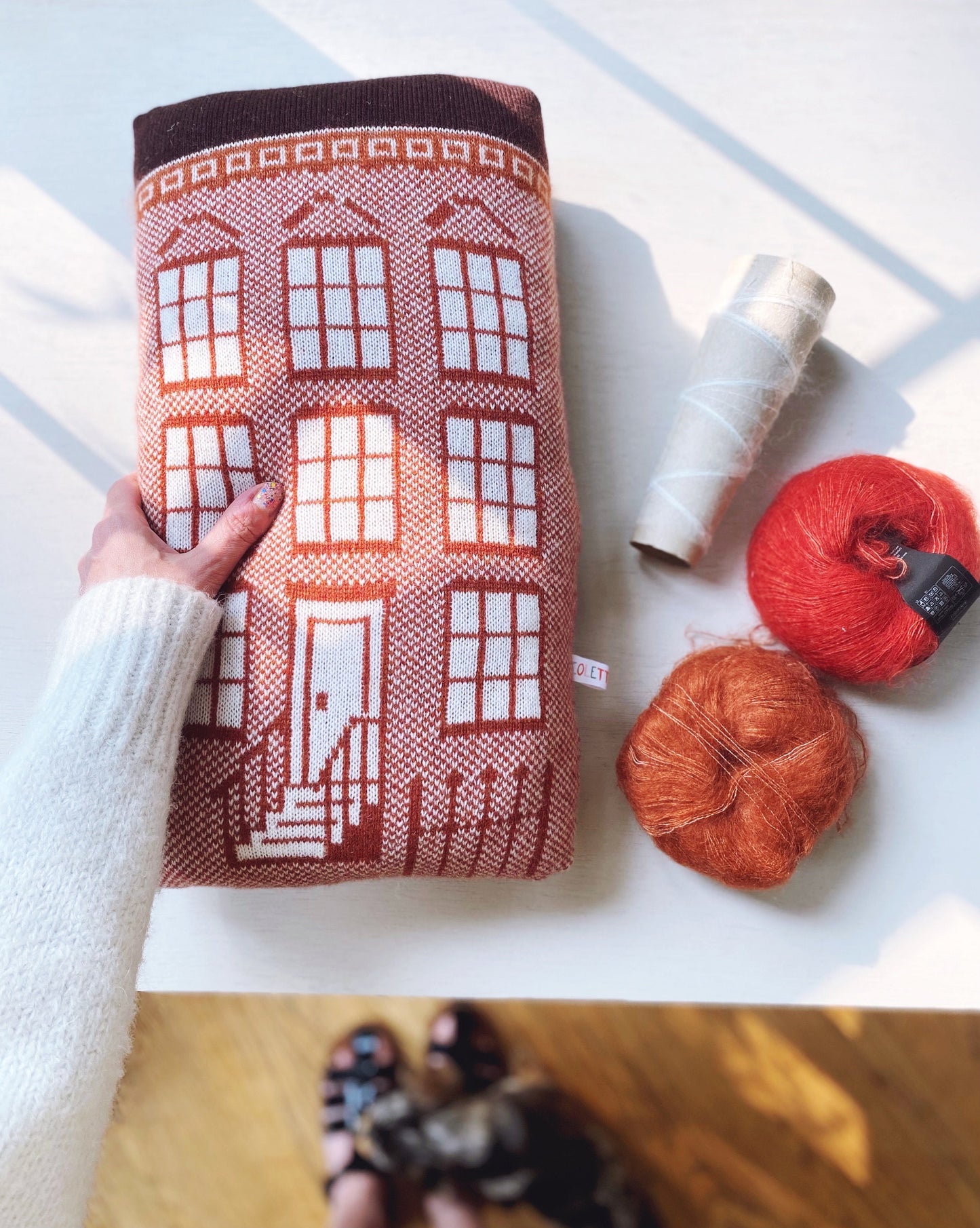 Knitted pillow with house pattern resembling a brownstone held by a hand, next to yarn on a light surface.