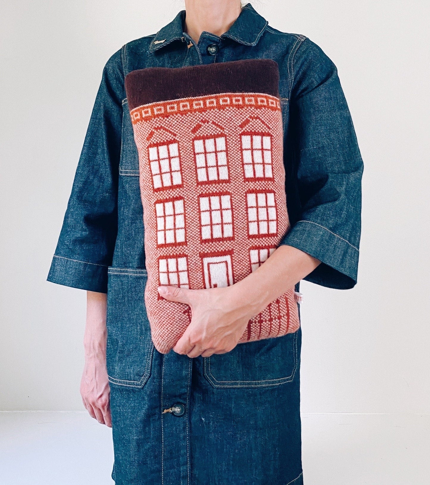 Person holding a brown house shaped knitted pillow resembling a brownstone against a plain background
