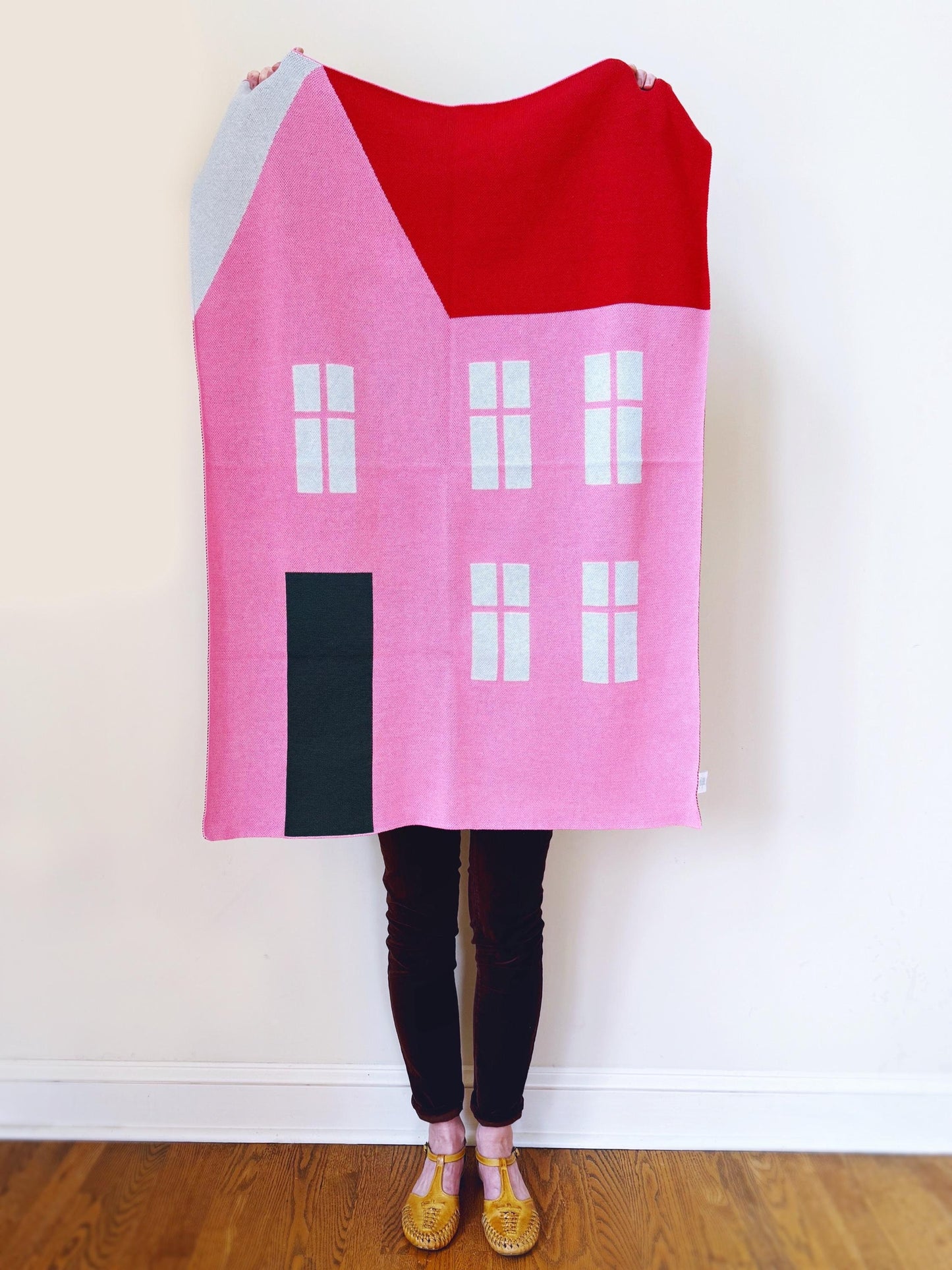 Person holding a pink house-shaped knitted blanket against a white wall.