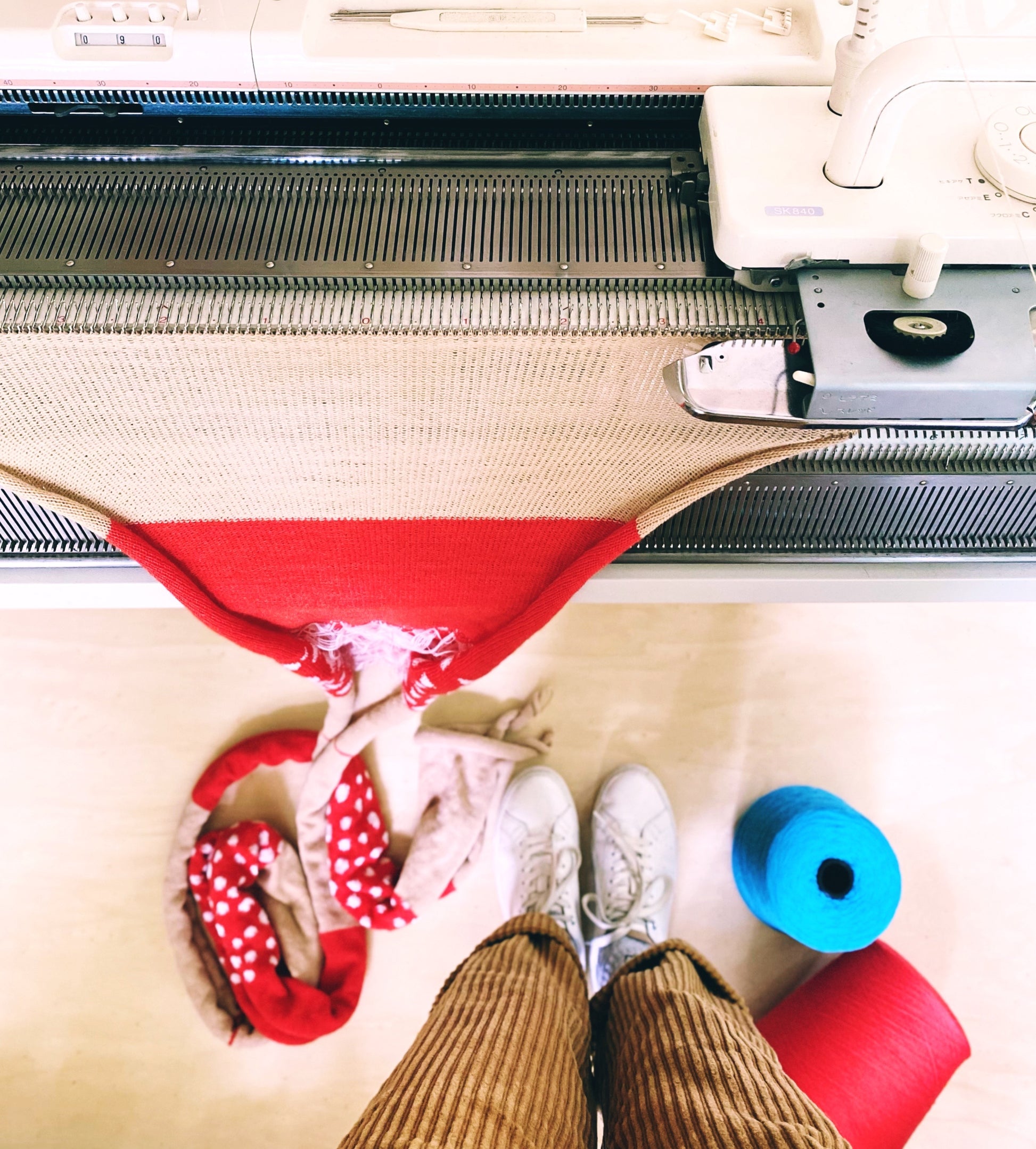 Person wearing white sneakers and brown pants standing next to a knitting machine with red fabric.