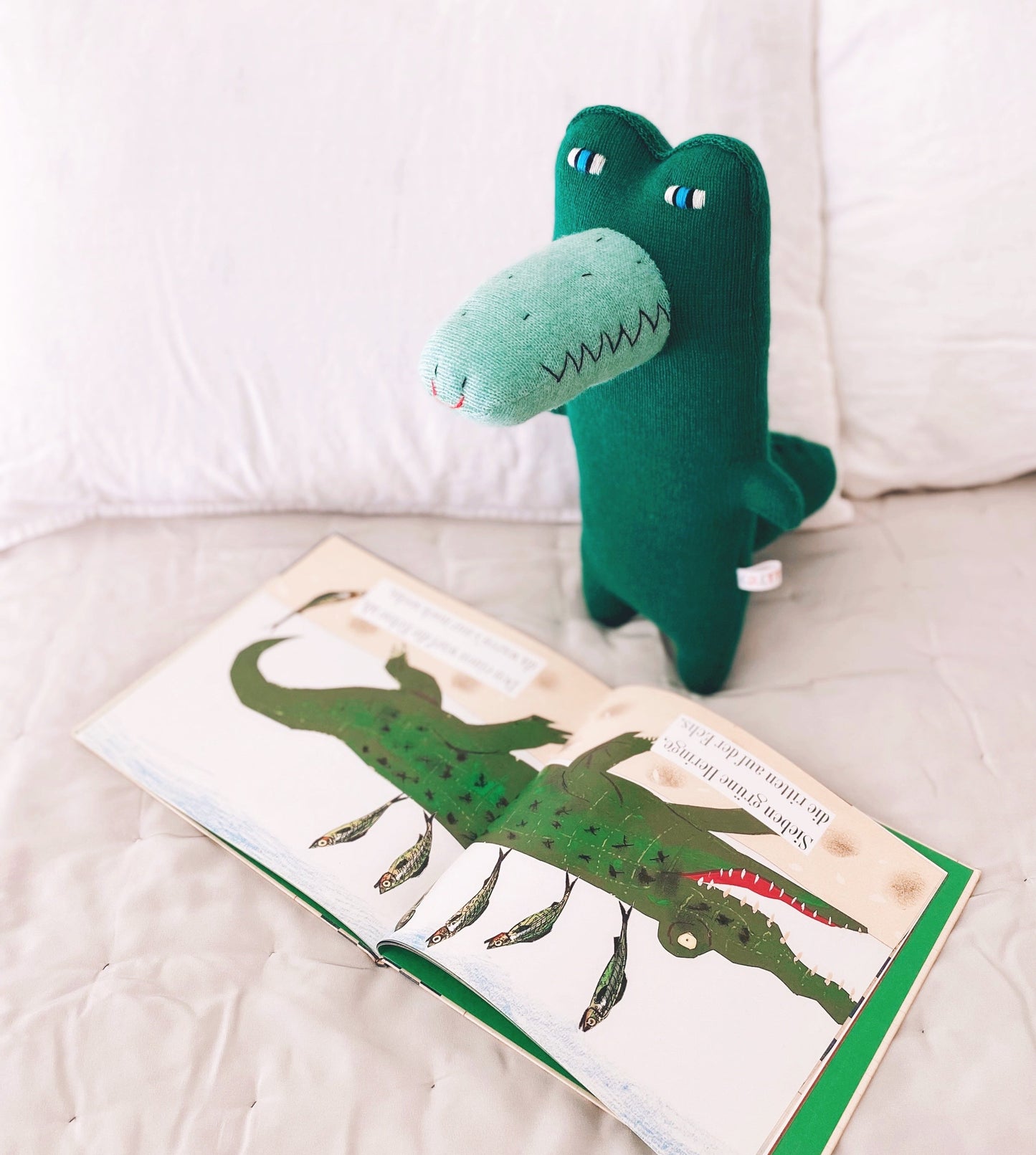 Green knitted lambswool plush toy alligator crocodile next to a book with an alligator illustration on a white surface.