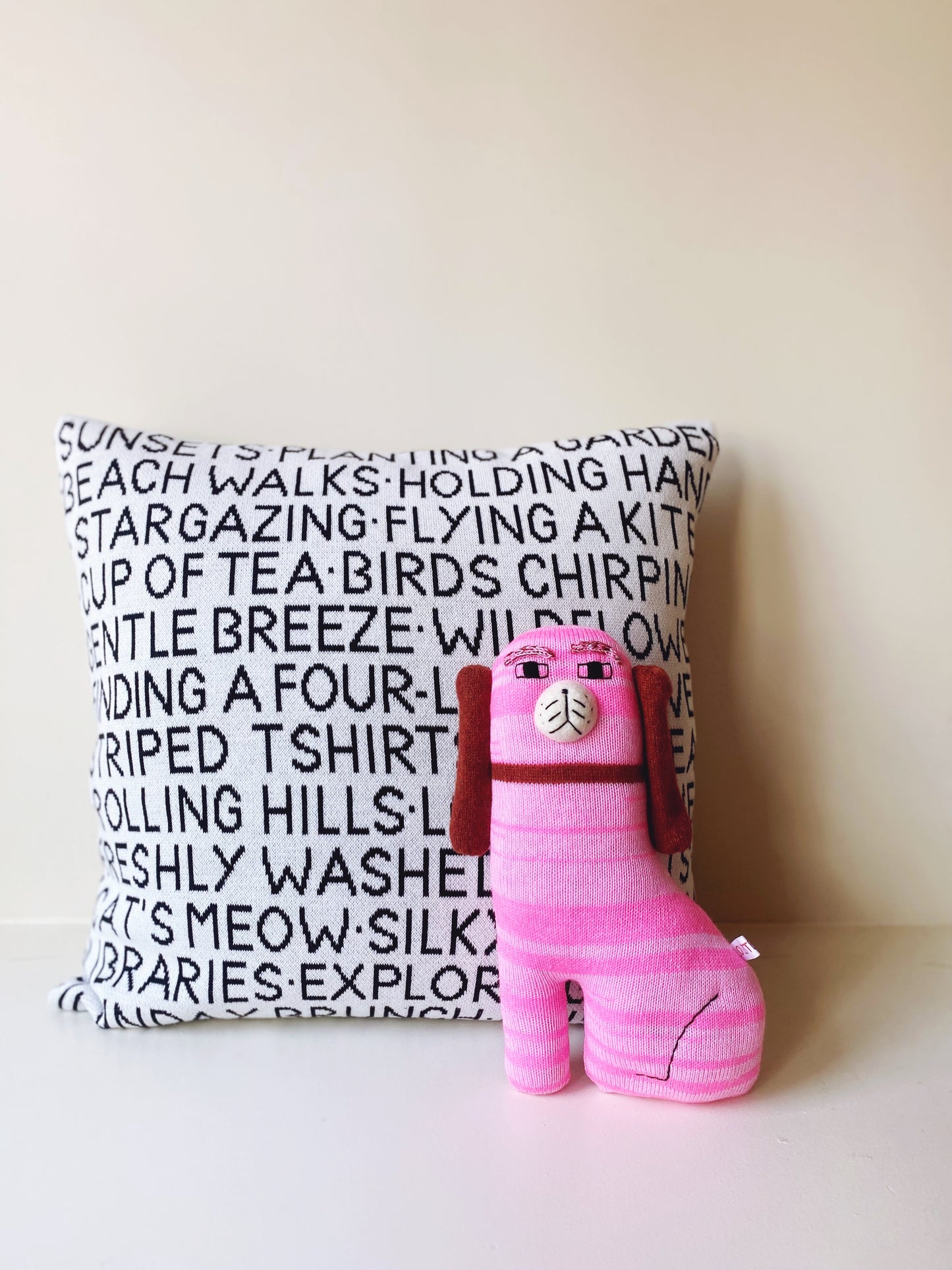 Pink knitted lambswool Staffordshire dog plush toy and a wordy knitted pillow with text on it on white background