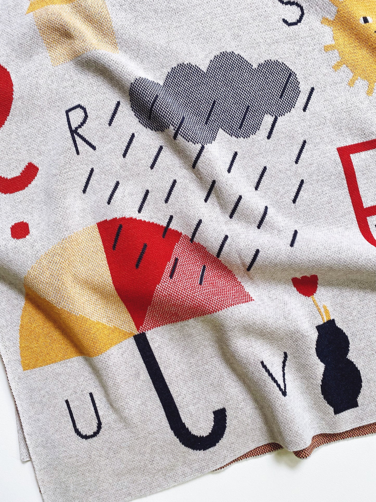 Knitted alphabet blanket throw with raindrop, umbrella, and sun designs on a light gray background