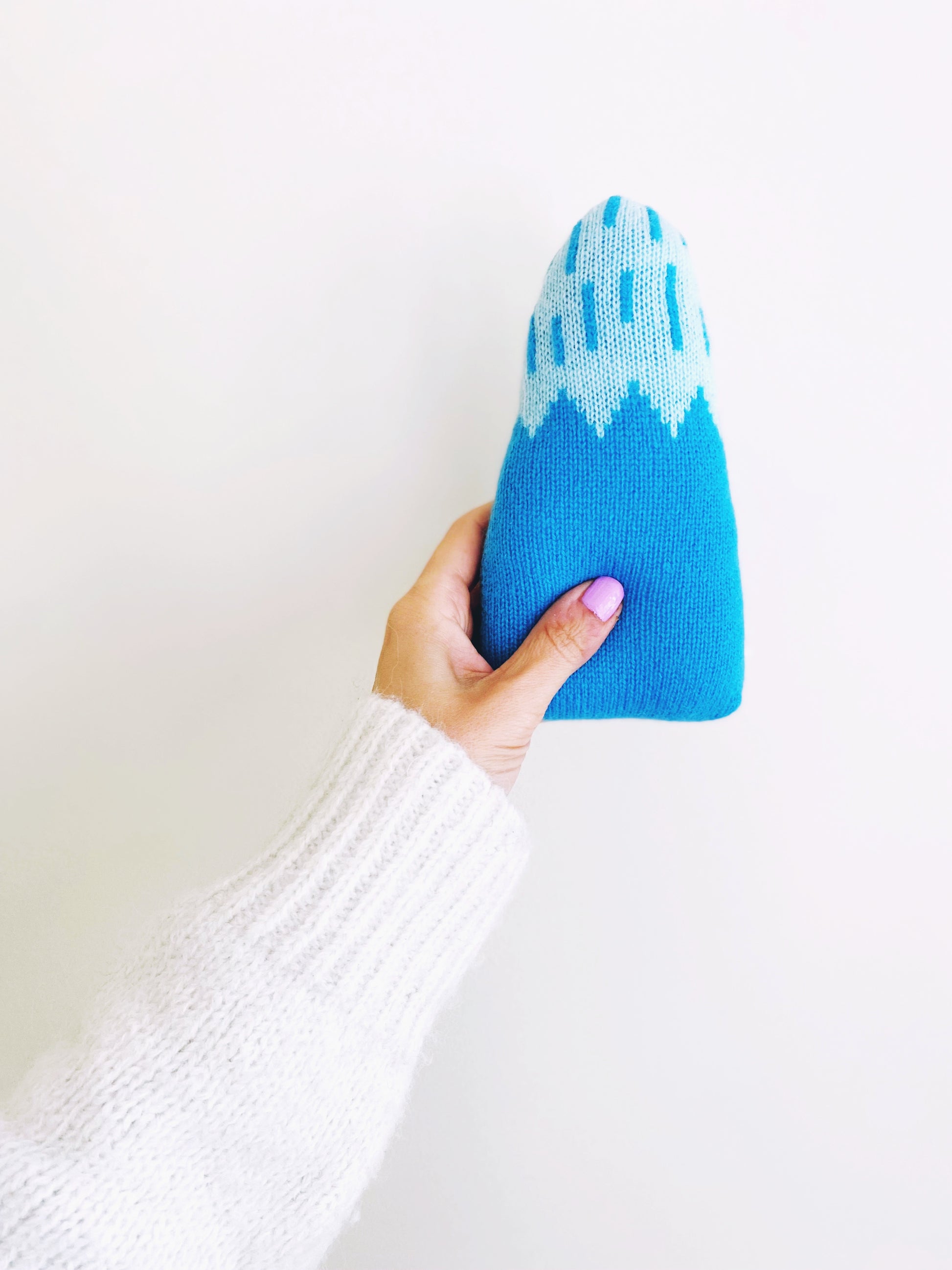 Hand holding a blue and white knitted baby rattle toy shaped like a mountain against a white background