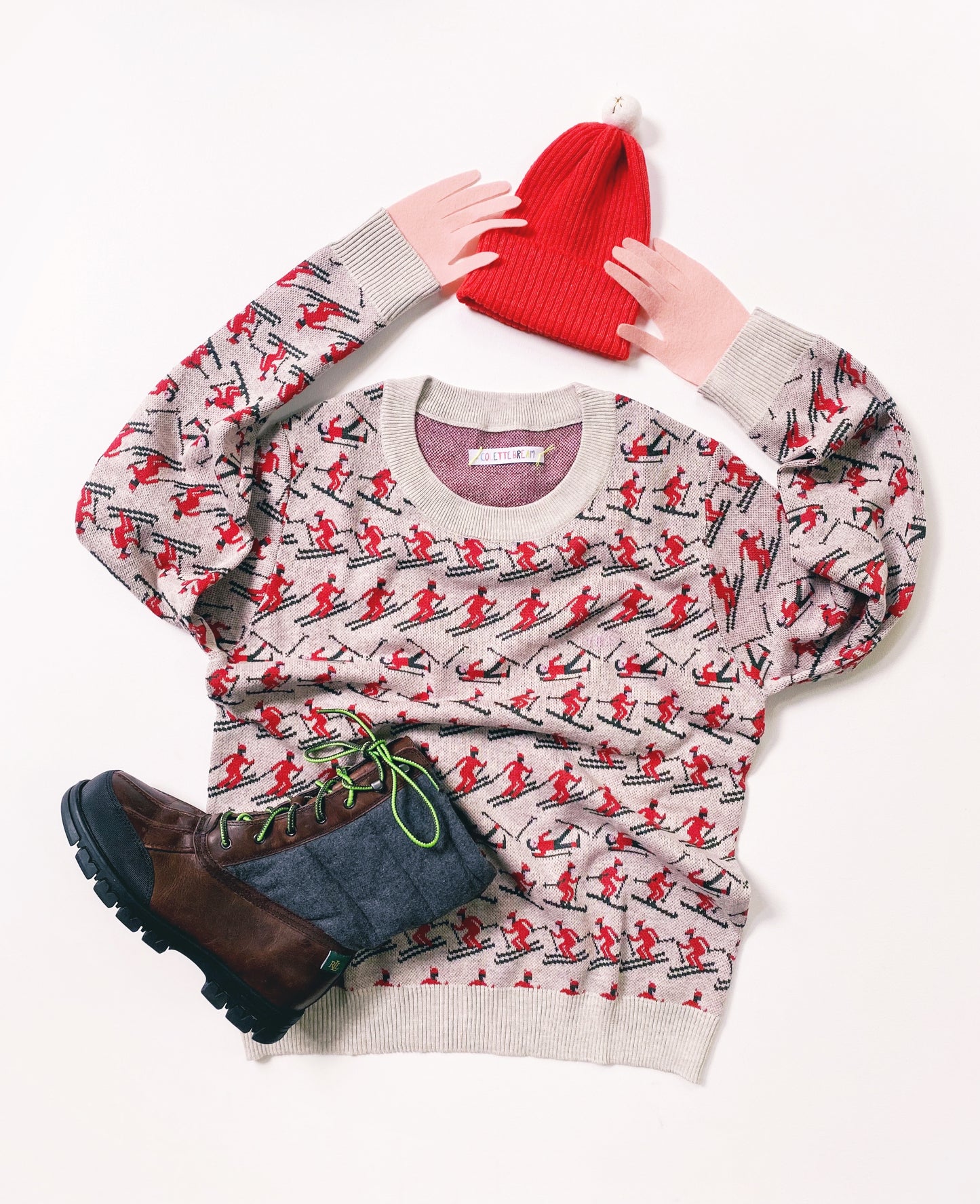 Knitted sweater with red and black pattern of skiers, red beanie, and brown boots on a white background