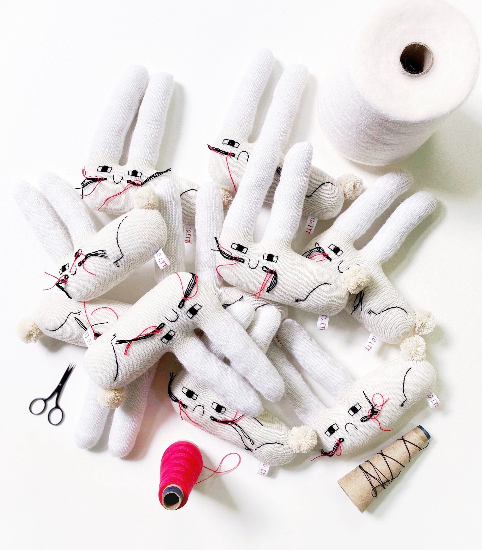 White plush toys with bunny rabbit faces on a white background, surrounded by spools of thread and scissors.