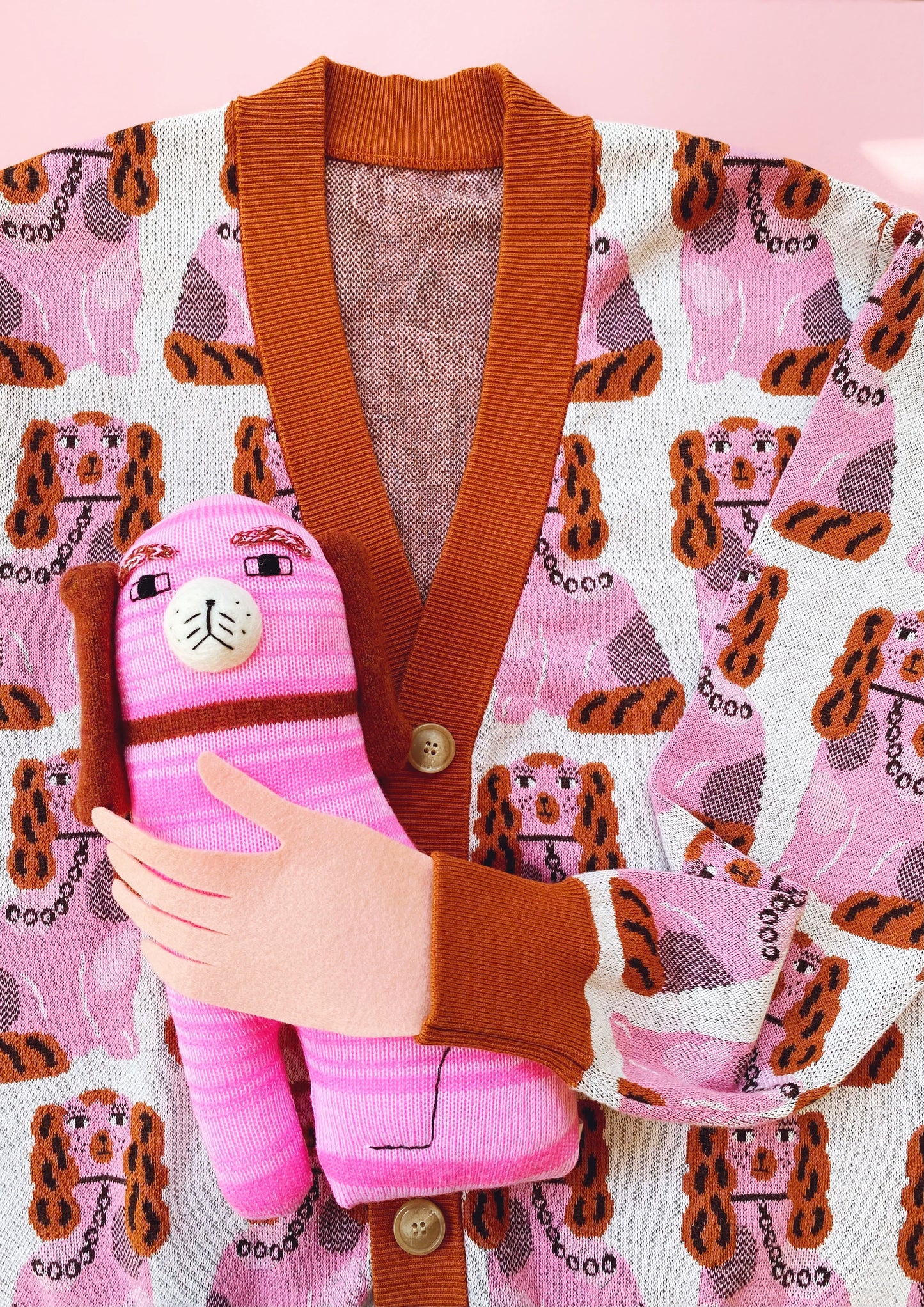 Pink Staffordshire dog plush toy held by a hand against a pink cardigan background with a Staffordshire dog pattern.