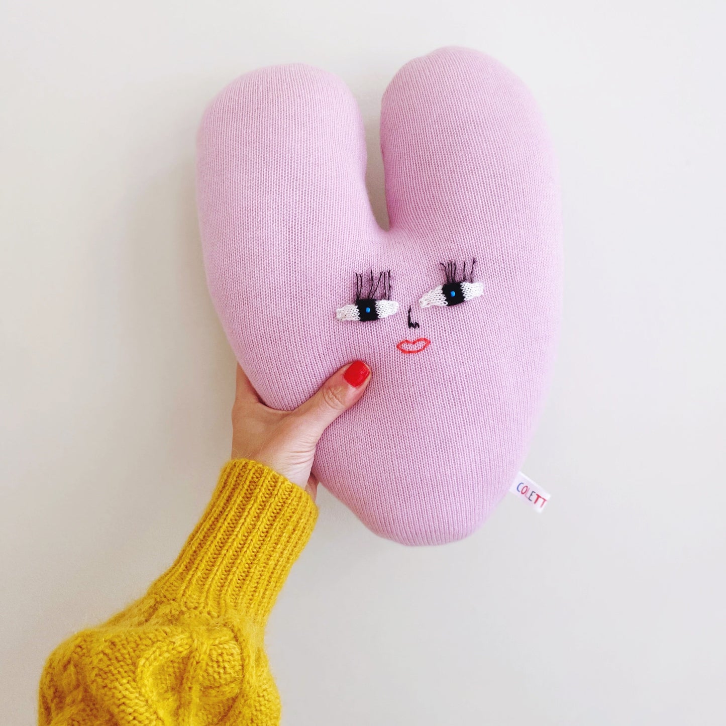 Pink knitted heart-shaped pillow with face design held by a person wearing a yellow sweater on a light background