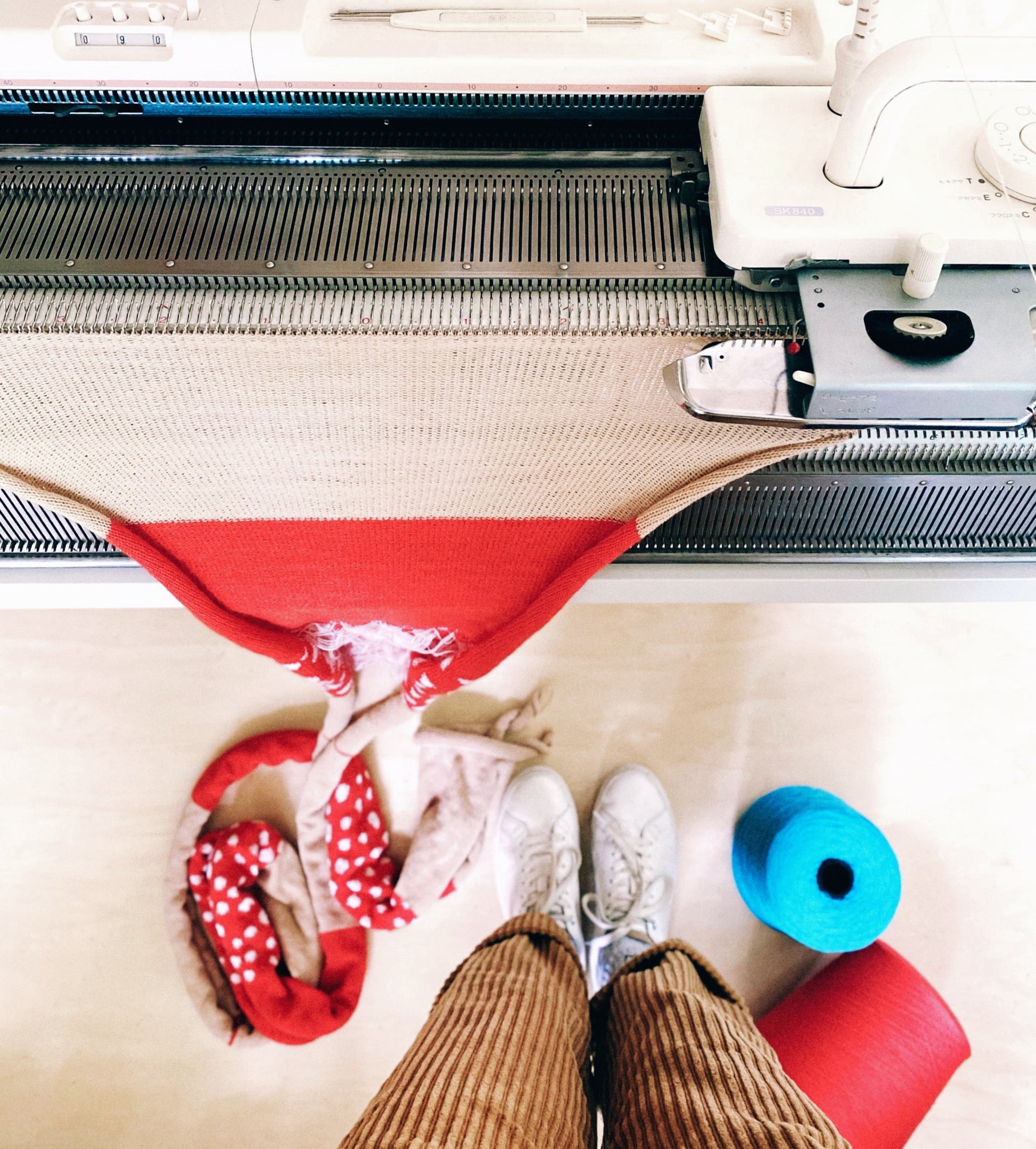 Person using a knitting machine with a red fabric and white shoes on a light surface.