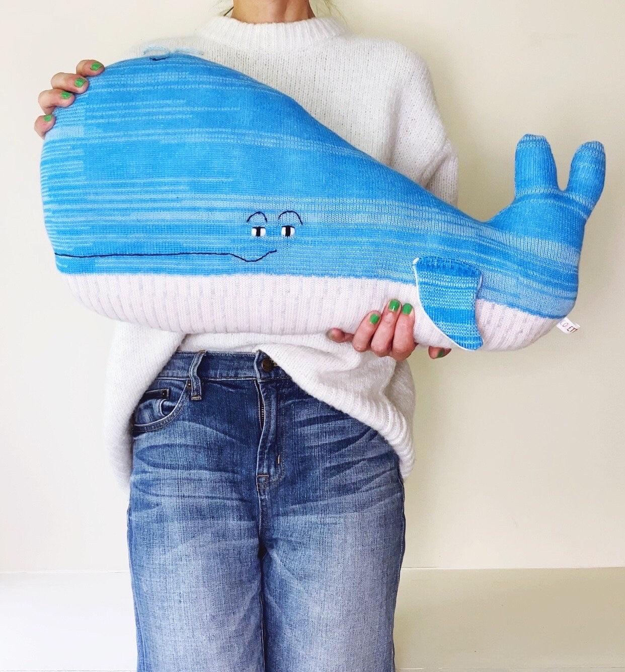 Person holding a blue lambswool knittedwhale-shaped pillow against a plain background
